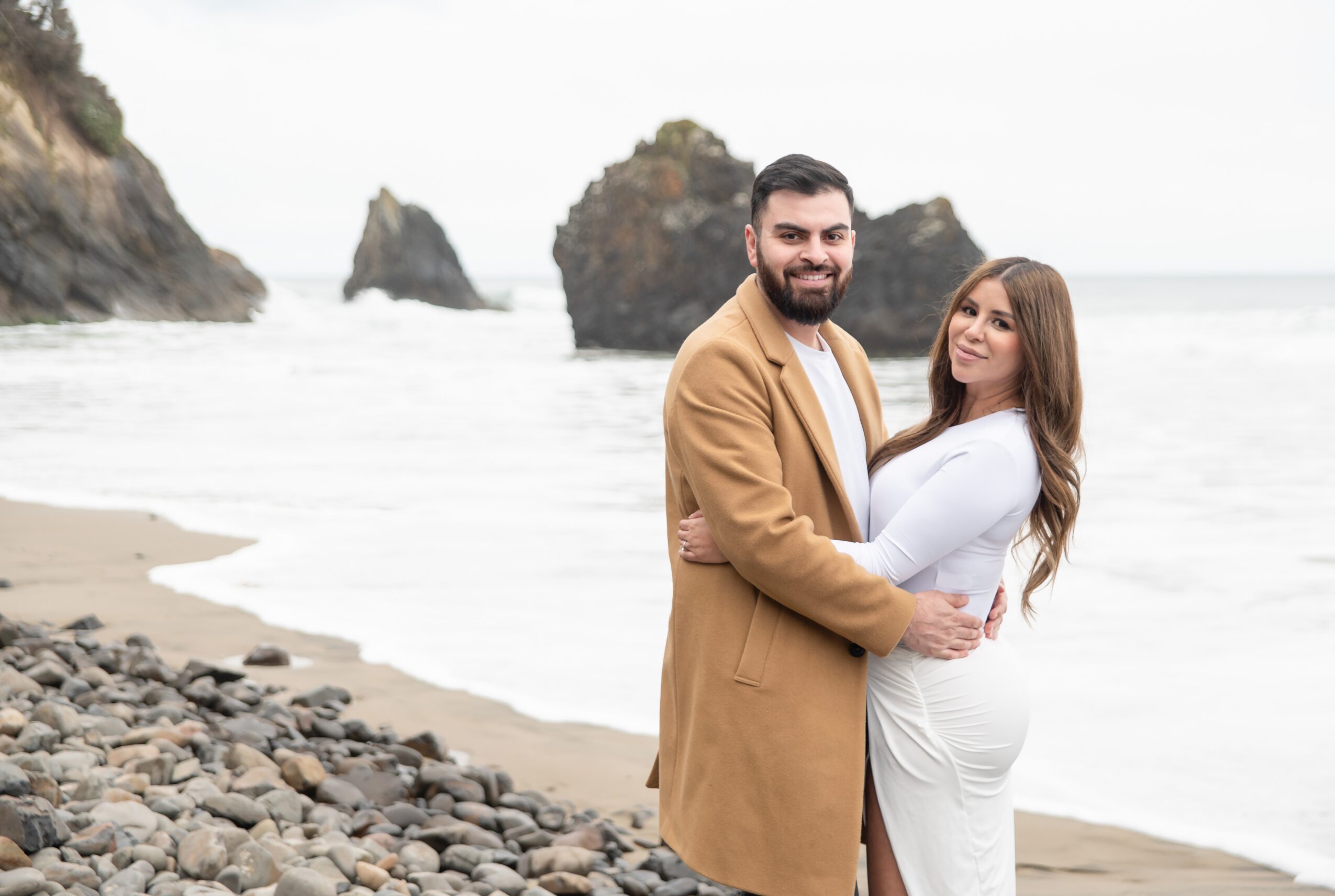 A smiling couple stands on a rocky beach with ocean waves and large rock formations in the background.