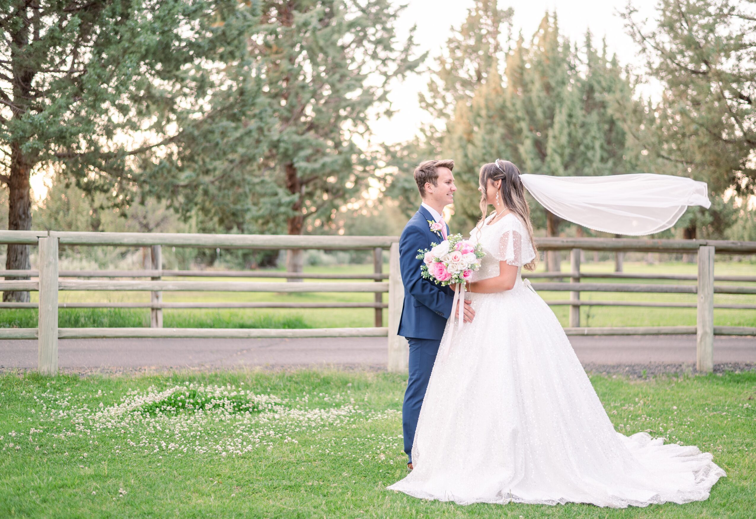 A bride and groom stand facing each other outdoors, holding a bouquet, with trees and a fence in the background.