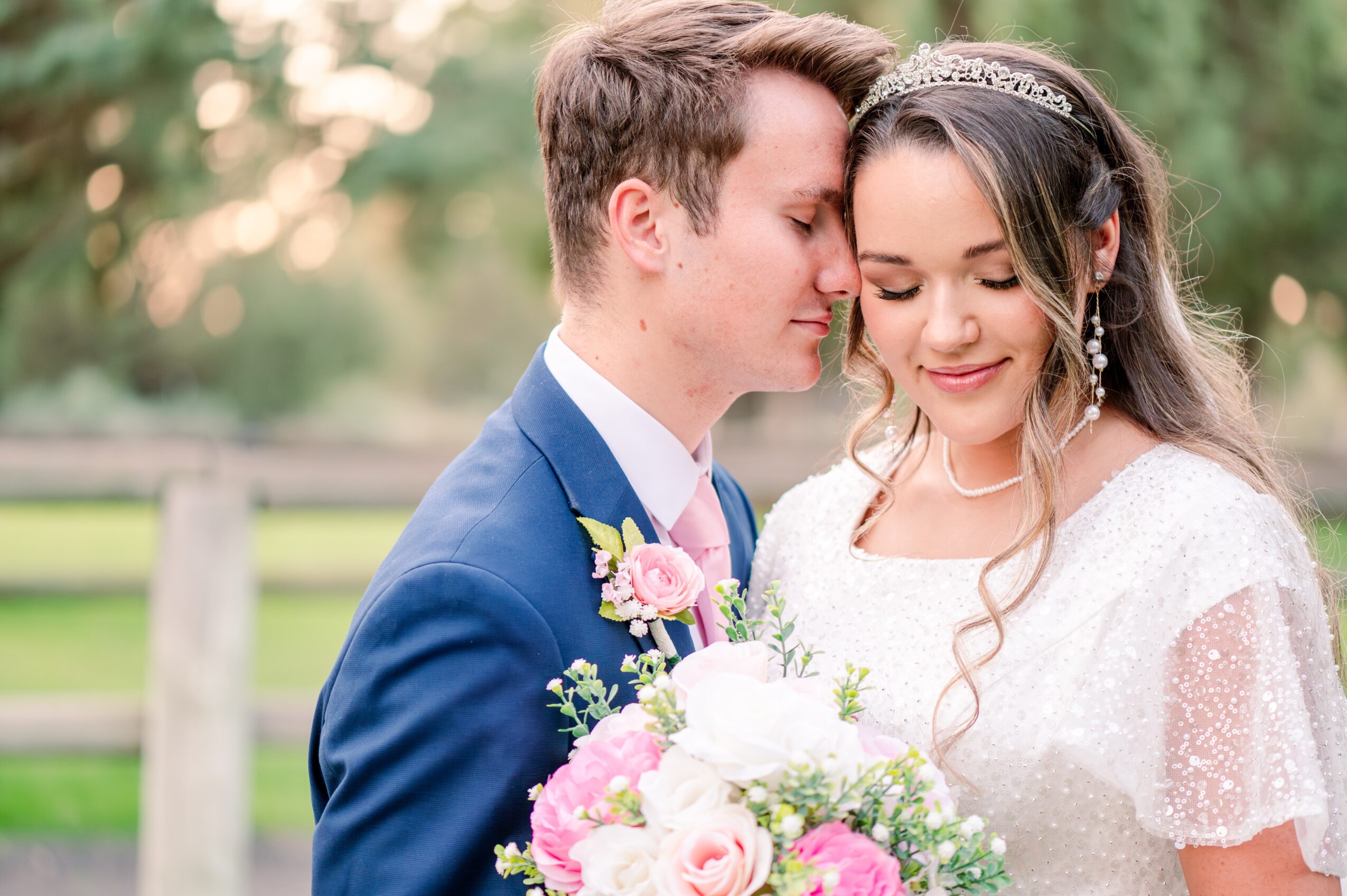 A couple in wedding attire close together outdoors, with trees and a fence in the background, holding a bouquet of flowers.