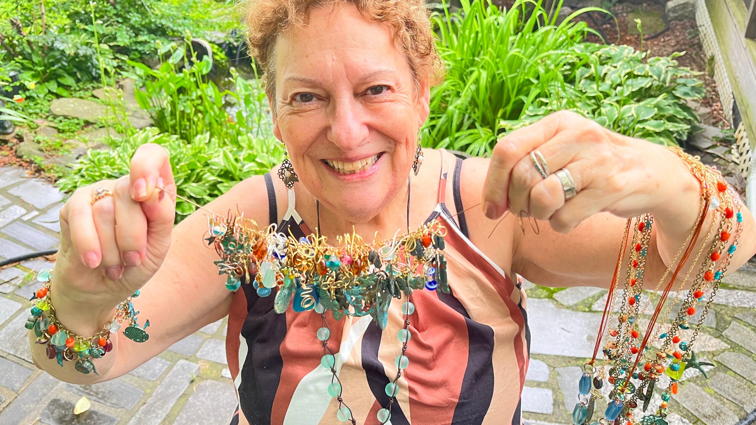 Smiling woman holding colorful jewelry outdoors with greenery in background.