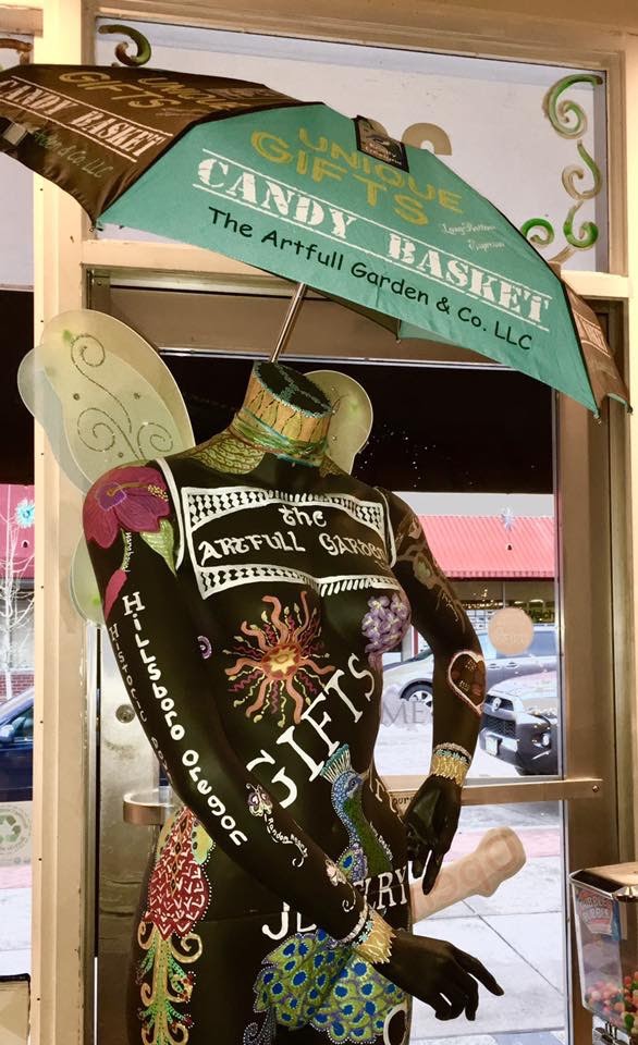 Display with a mannequin wearing a black shirt, holding a sign, and surrounded by decorative items, inside a glass window.