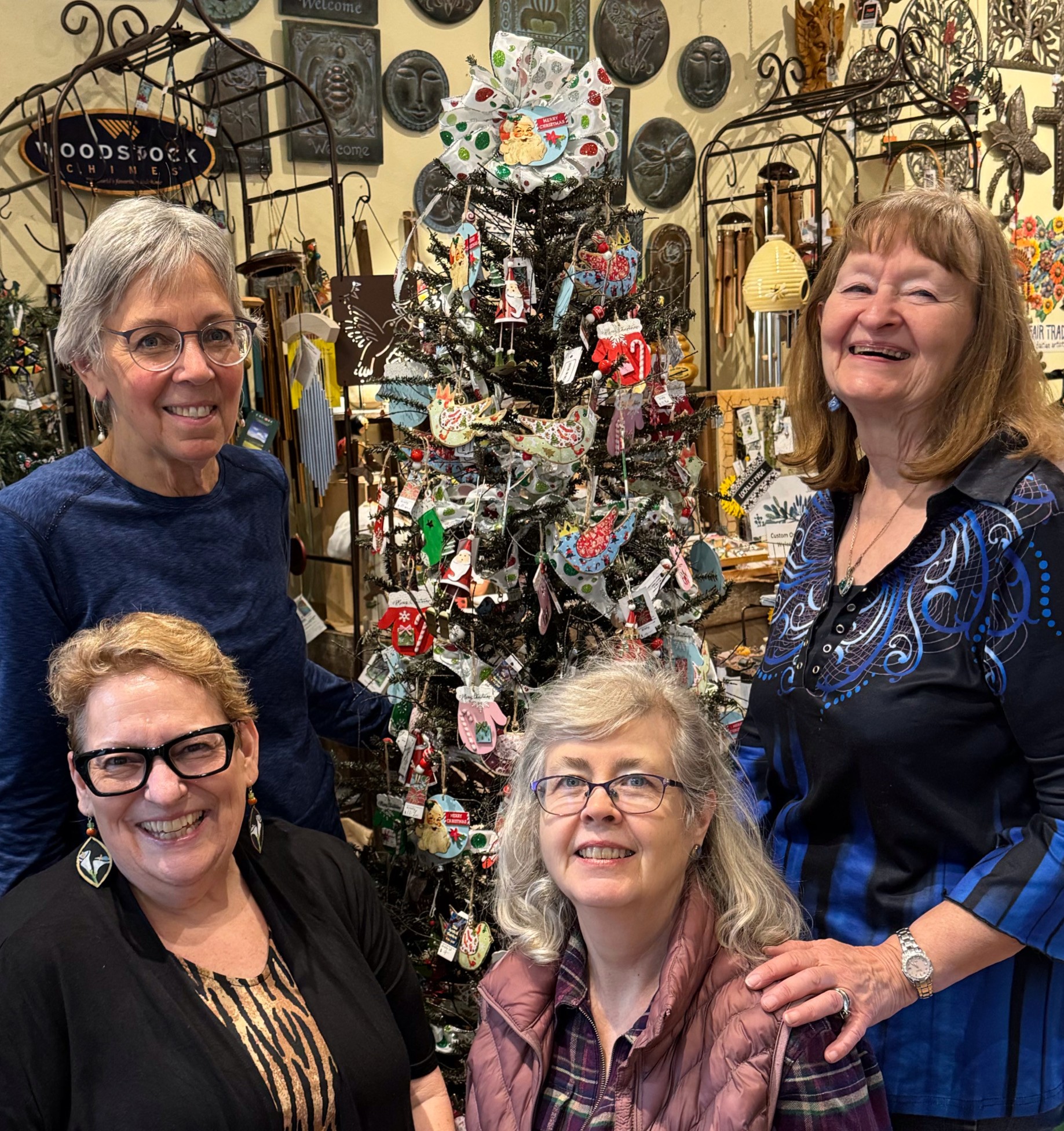 Four women smiling in front of a decorated Christmas tree with ornaments and artwork in the background.