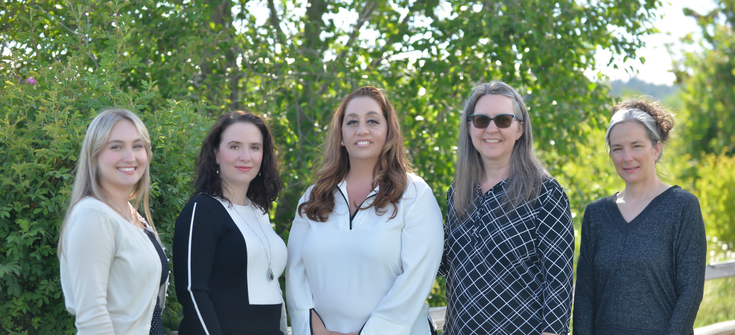 Six women standing outdoors in front of green trees, smiling, with sunlight filtering through leaves.