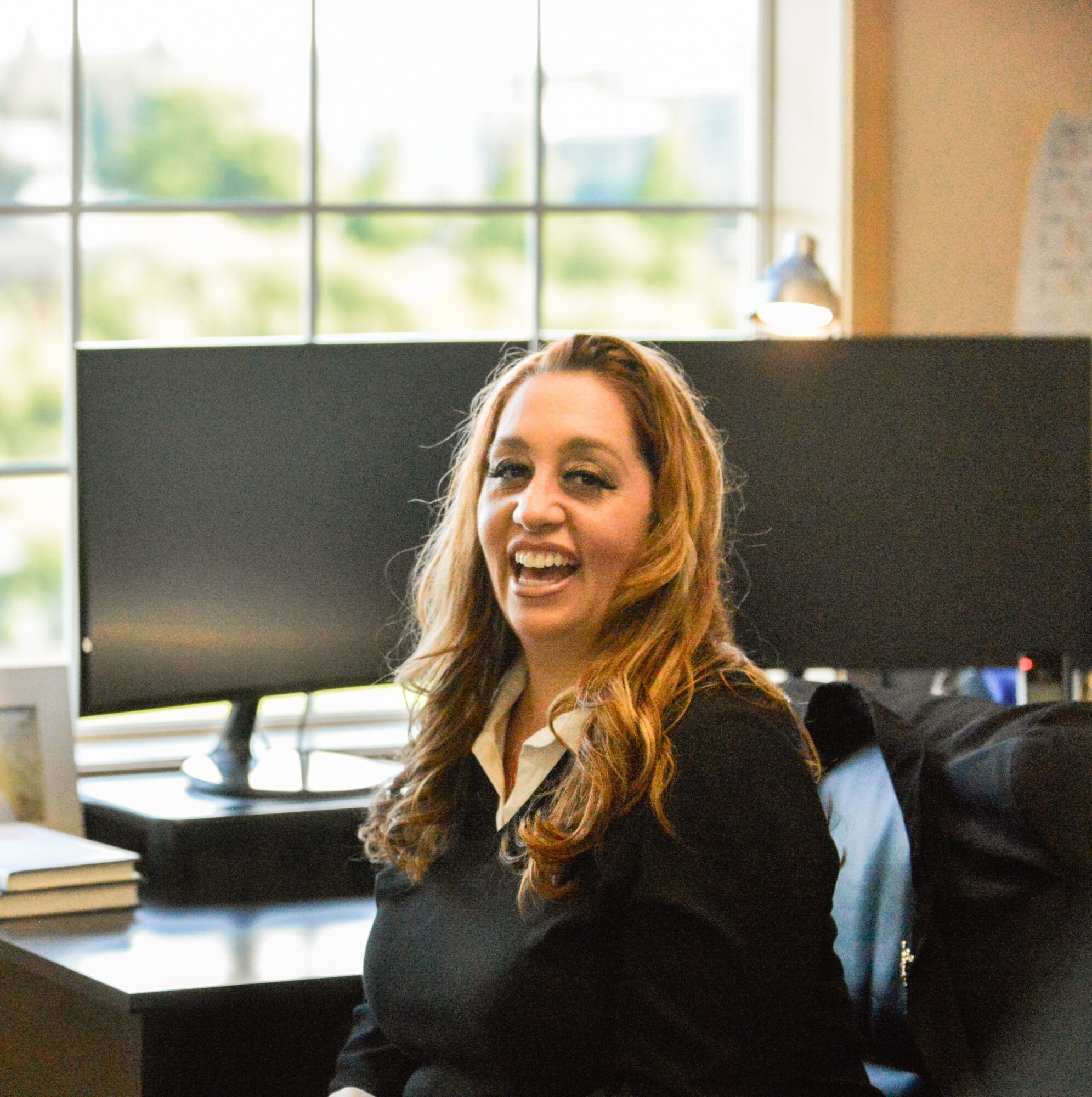 Young woman with long wavy hair smiling and laughing indoors near a window, with a desk and computer behind her.