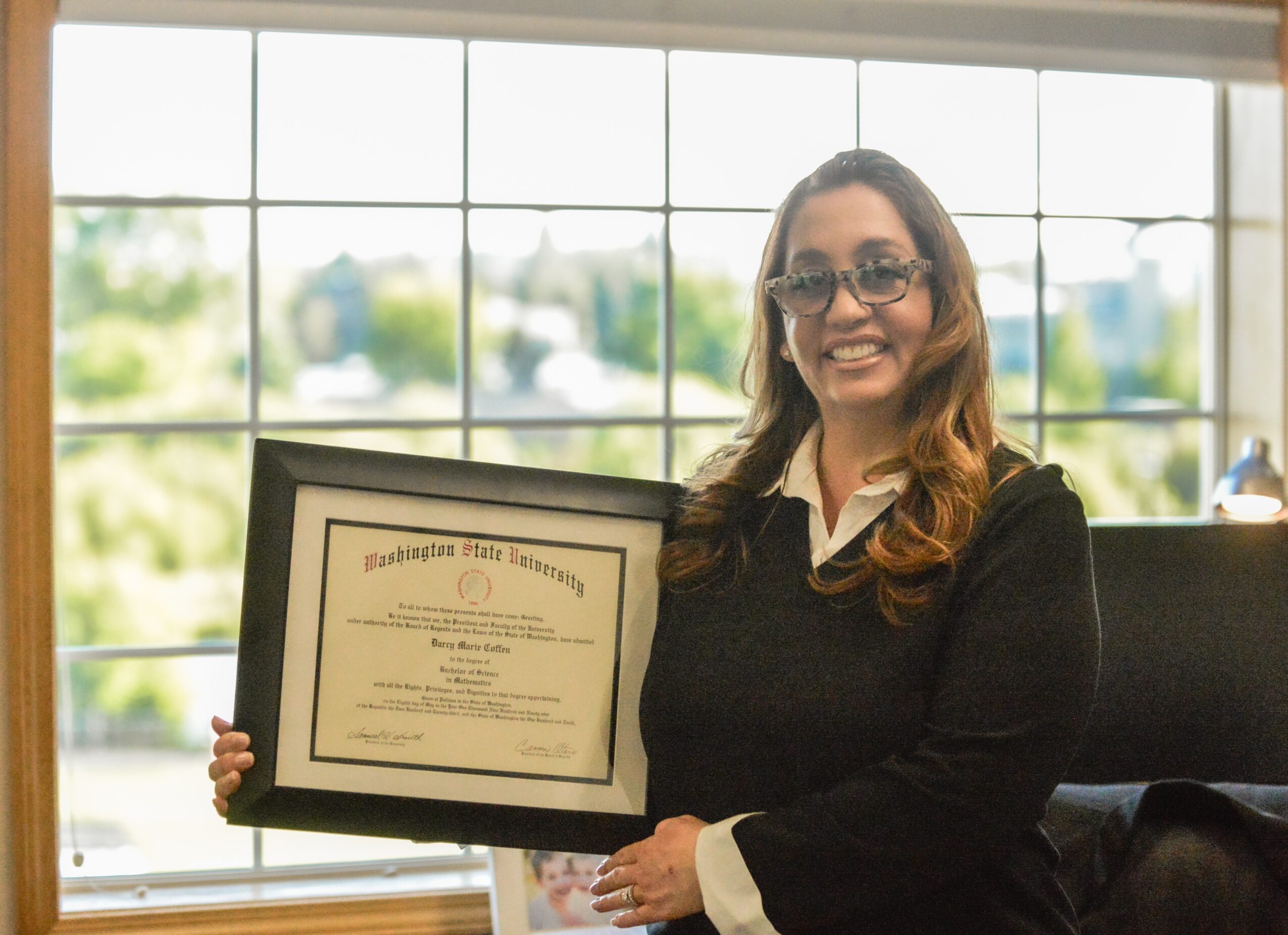 Woman with glasses holding a framed certificate, smiling, in front of a large window with a view outside.
