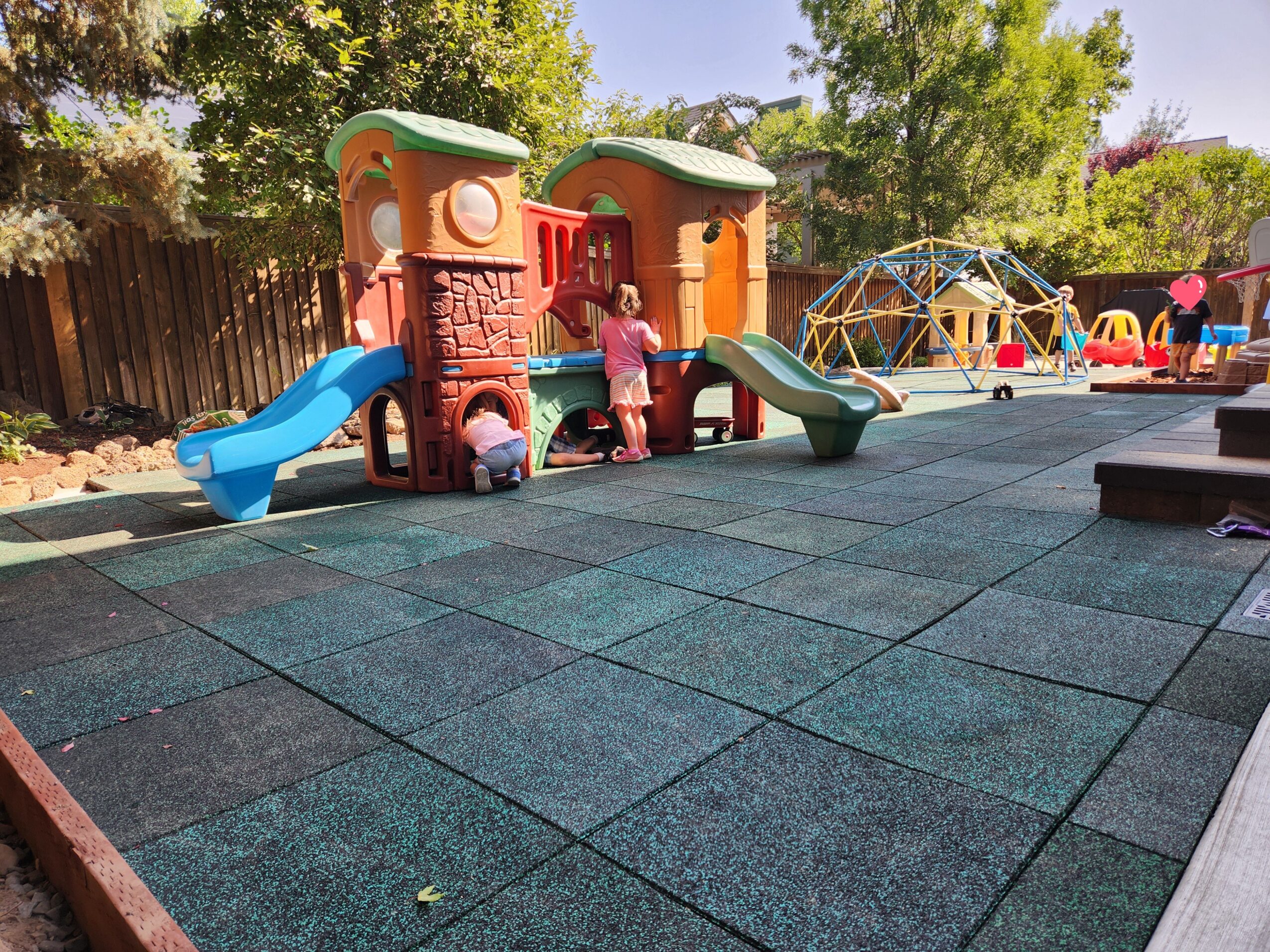 Playground with colorful slides, climbing structures, and a geodesic dome, surrounded by trees and a wooden fence.