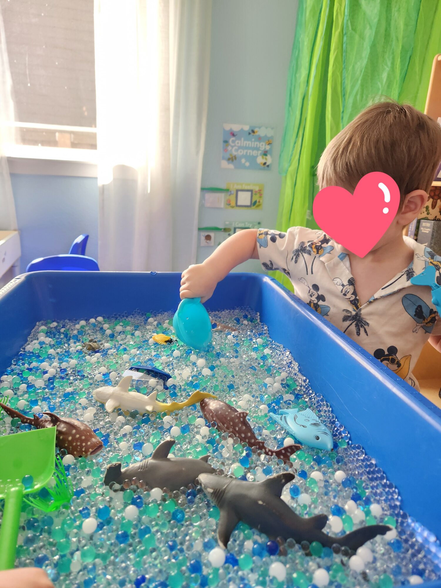Child playing with toy sharks in a water table filled with colorful beads, with a green curtain and window in background.