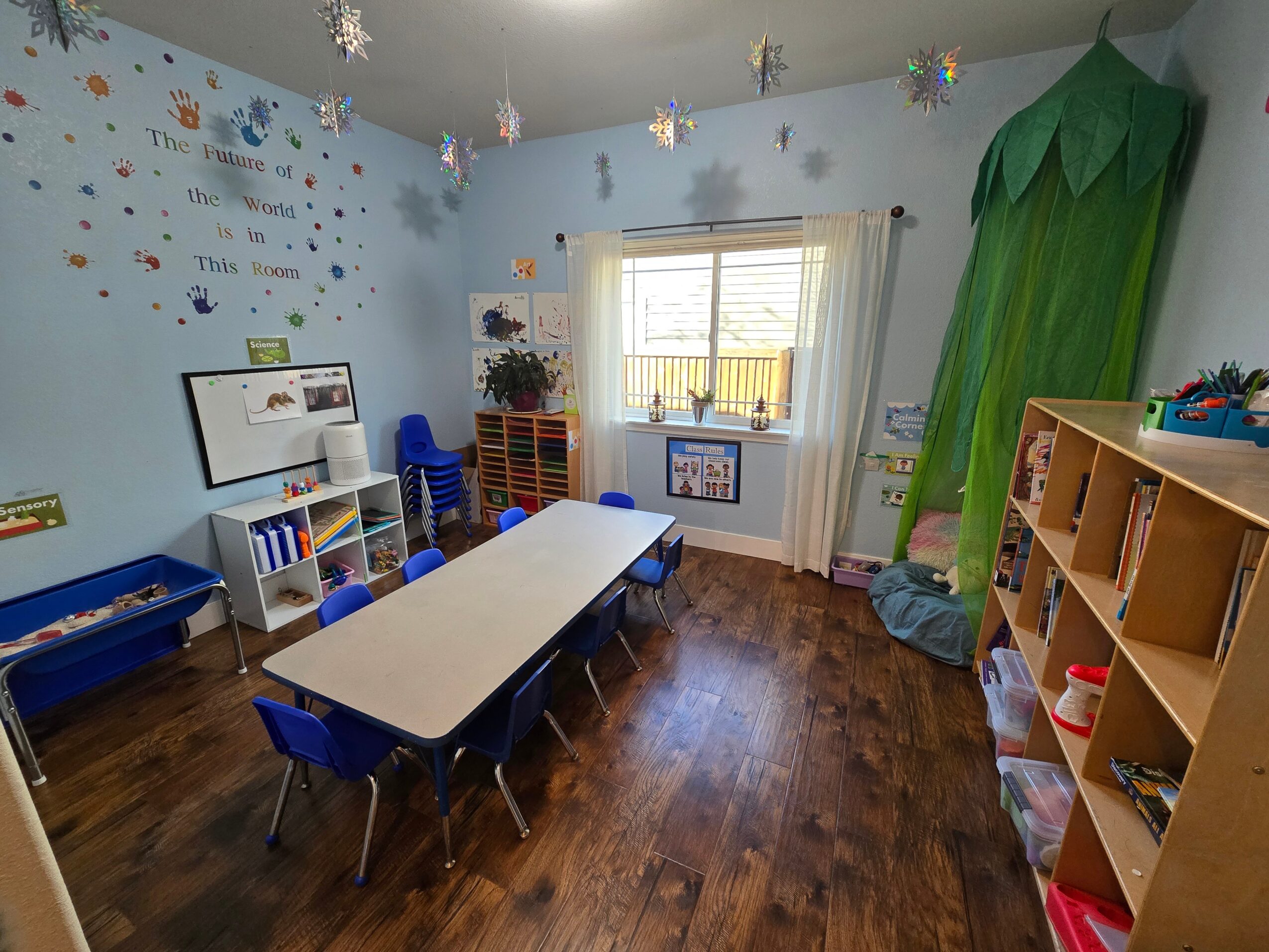 Classroom with a table, chairs, bookshelf, window, and green paper tree, decorated with stars and wall stickers.