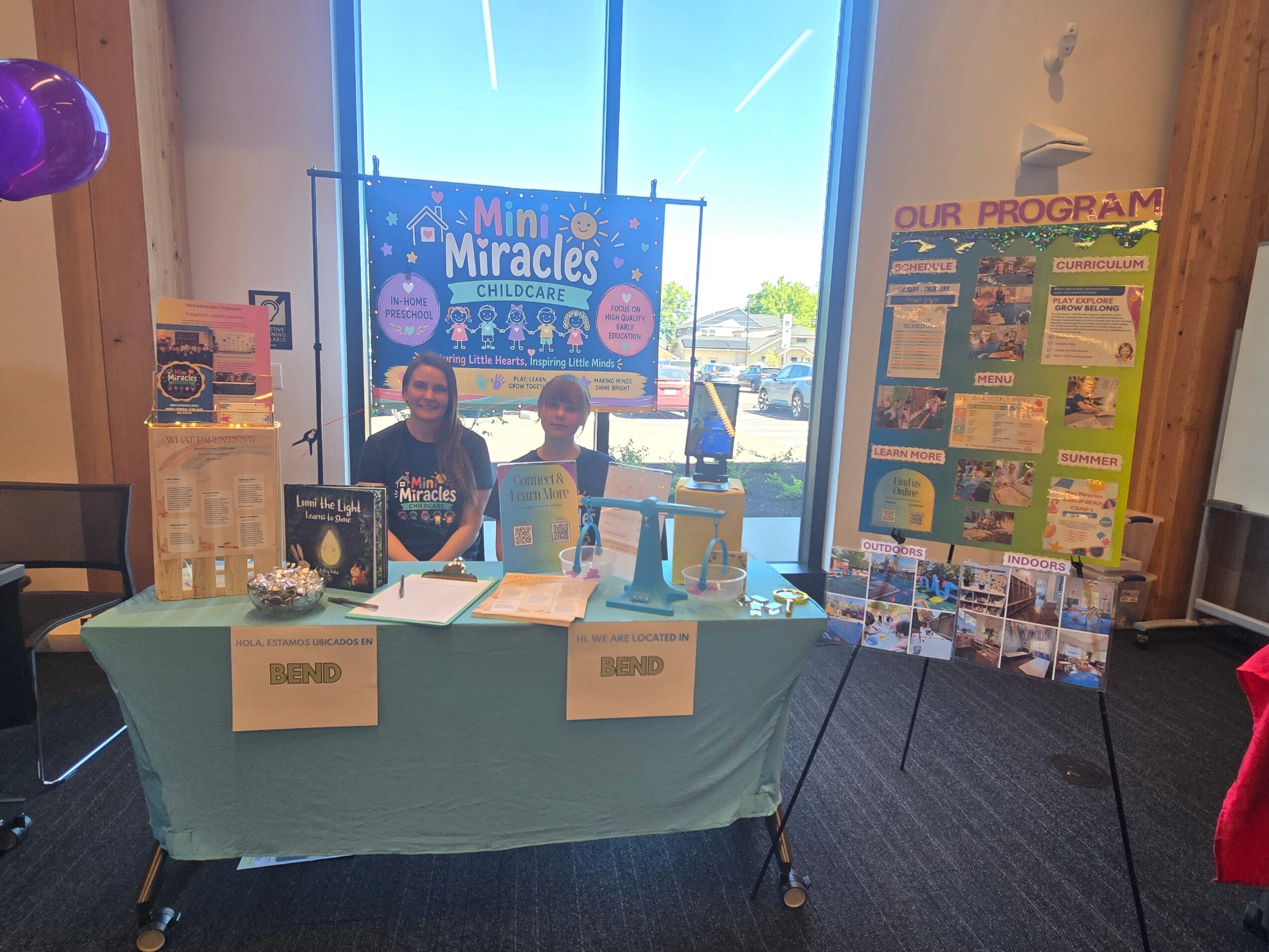 Two children stand behind a table with informational materials, in front of a window with a colorful banner reading 'Mini Miracles'.