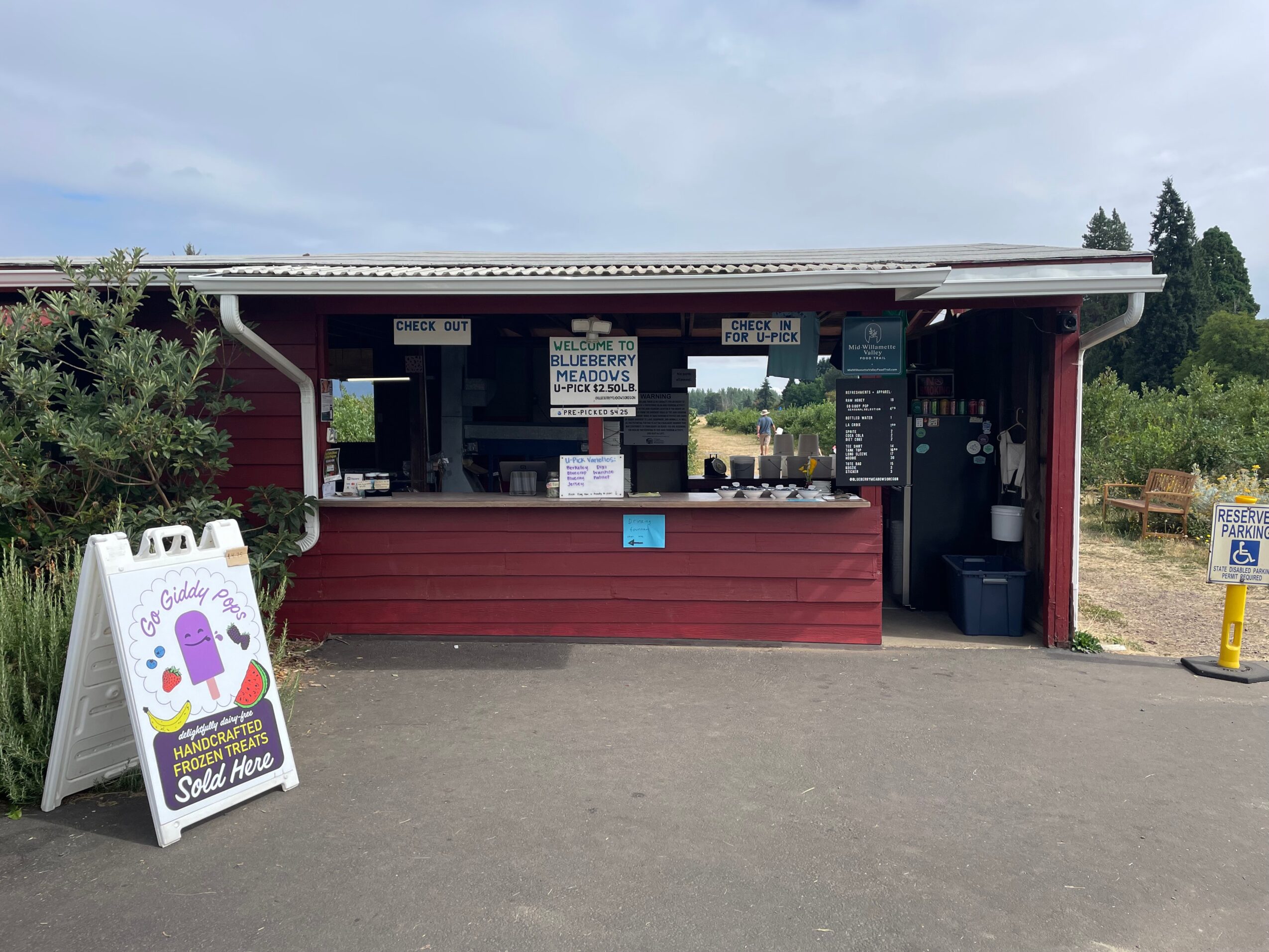 Small food stand with a red exterior, menu signs, and a white sandwich board outside, under a cloudy sky.