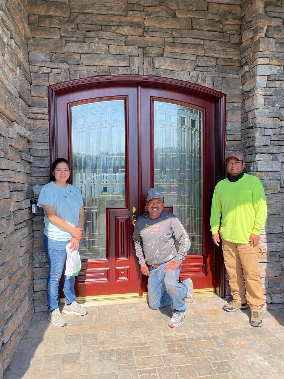 Three people standing in front of a large arched glass door with a brick wall background.