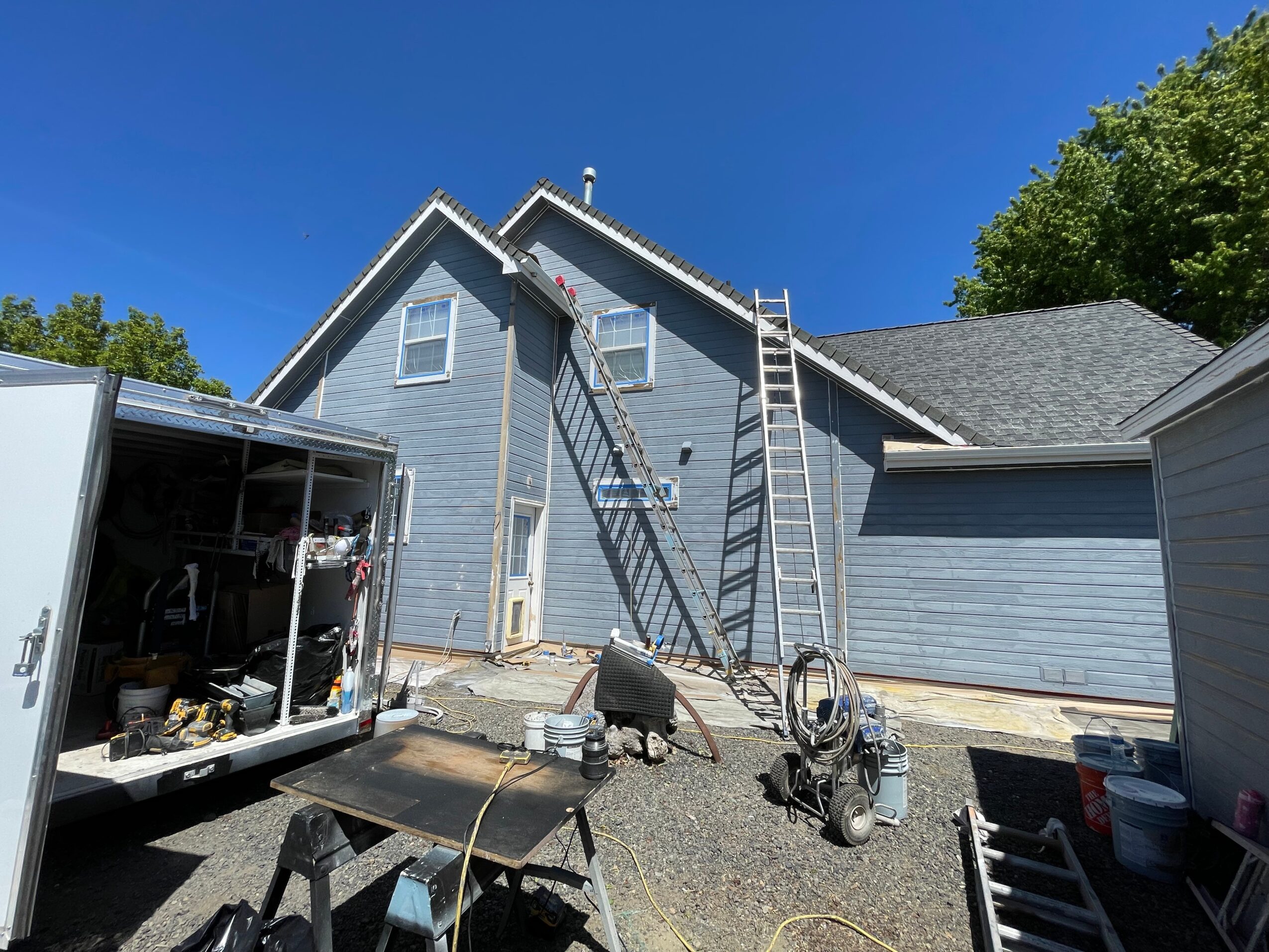 Side view of a blue house with a ladder leaning against it, construction equipment nearby, and a clear blue sky.