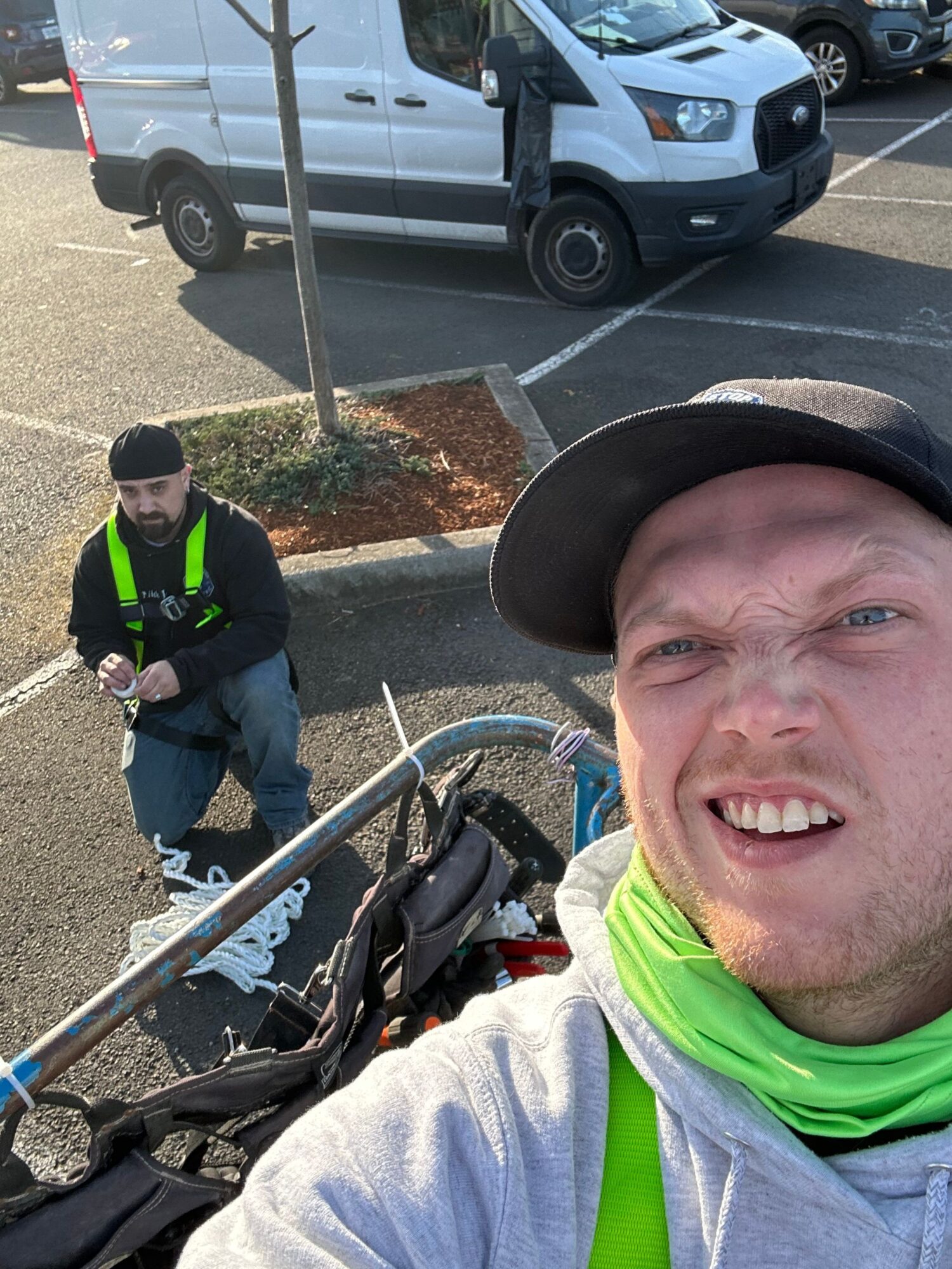 Person taking a selfie outdoors with a man in the background, a car, and a parking lot. The person is smiling and wearing a hat.