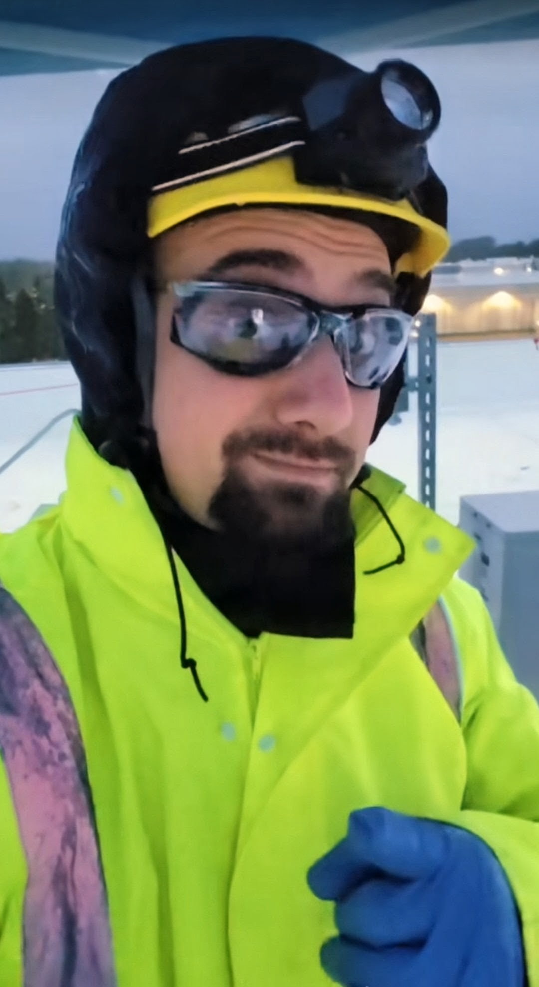 Person wearing safety gear, sunglasses, and a yellow jacket outdoors in snowy environment.