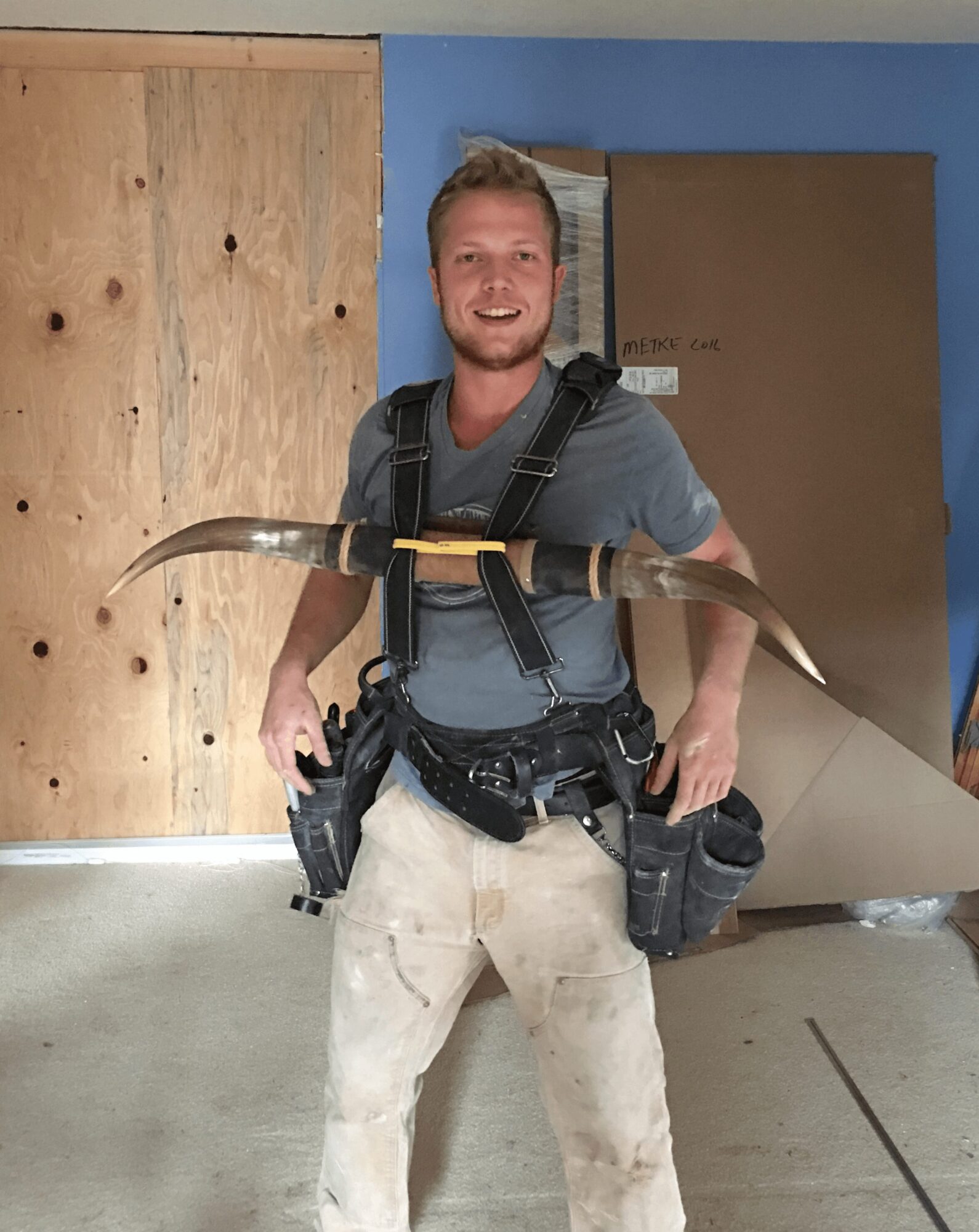 Young man with construction tools, smiling, standing indoors with a blue wall and wooden panel behind him.
