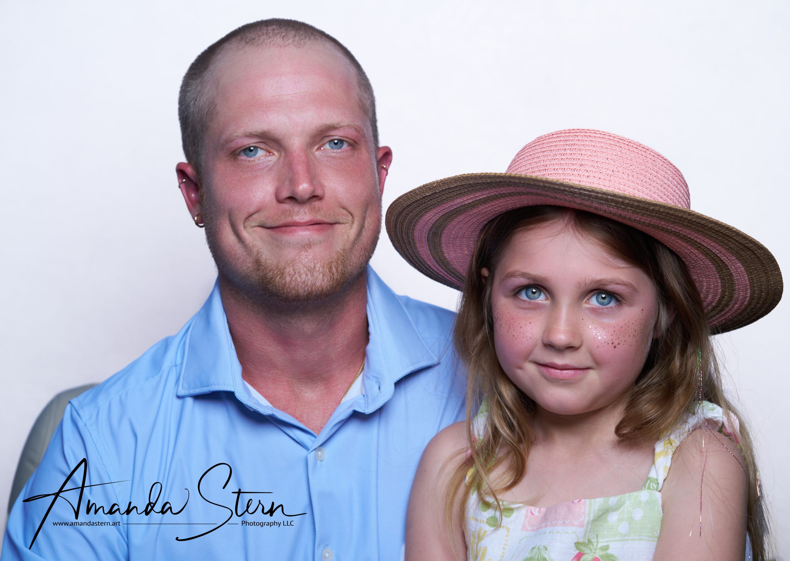 A man and a young girl pose together, both smiling, with the girl wearing a large pink hat and a light dress.