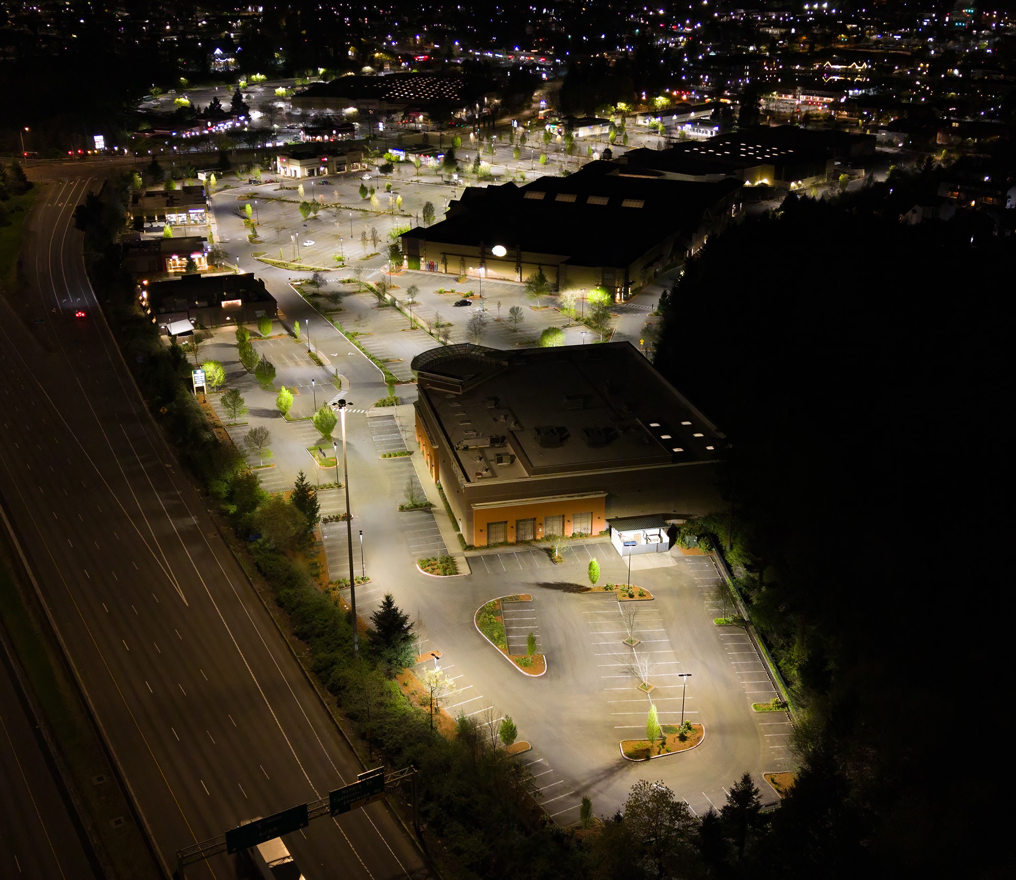 Nighttime aerial view of a parking lot with illuminated trees and buildings, adjacent to a highway on the left.