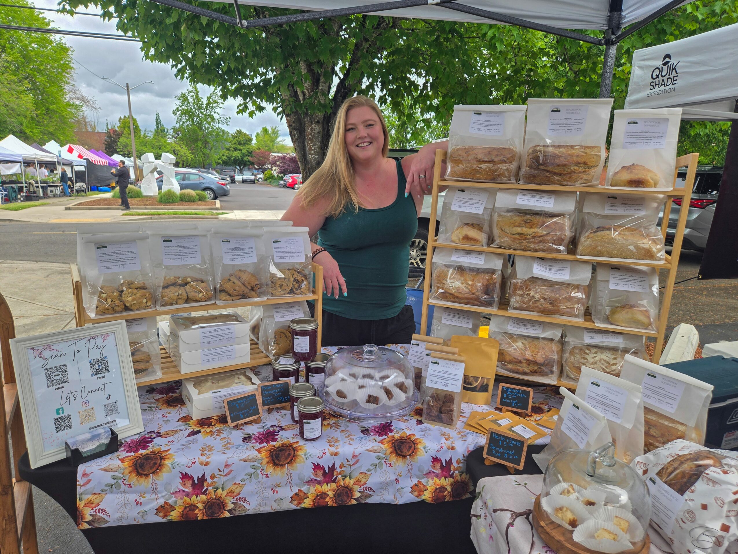 Smiling woman standing behind a table with baked goods at an outdoor market, trees and tents in background.