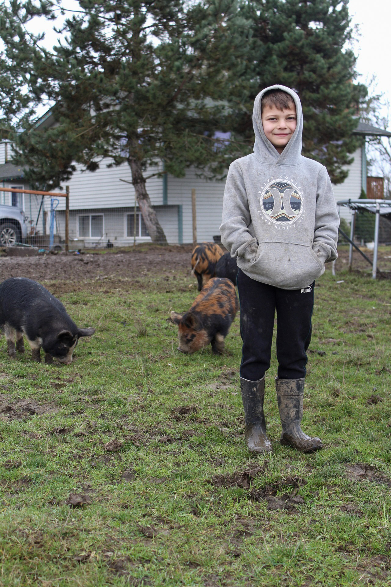 Boy in hoodie and boots standing outdoors with two dogs, trees, and houses in background.