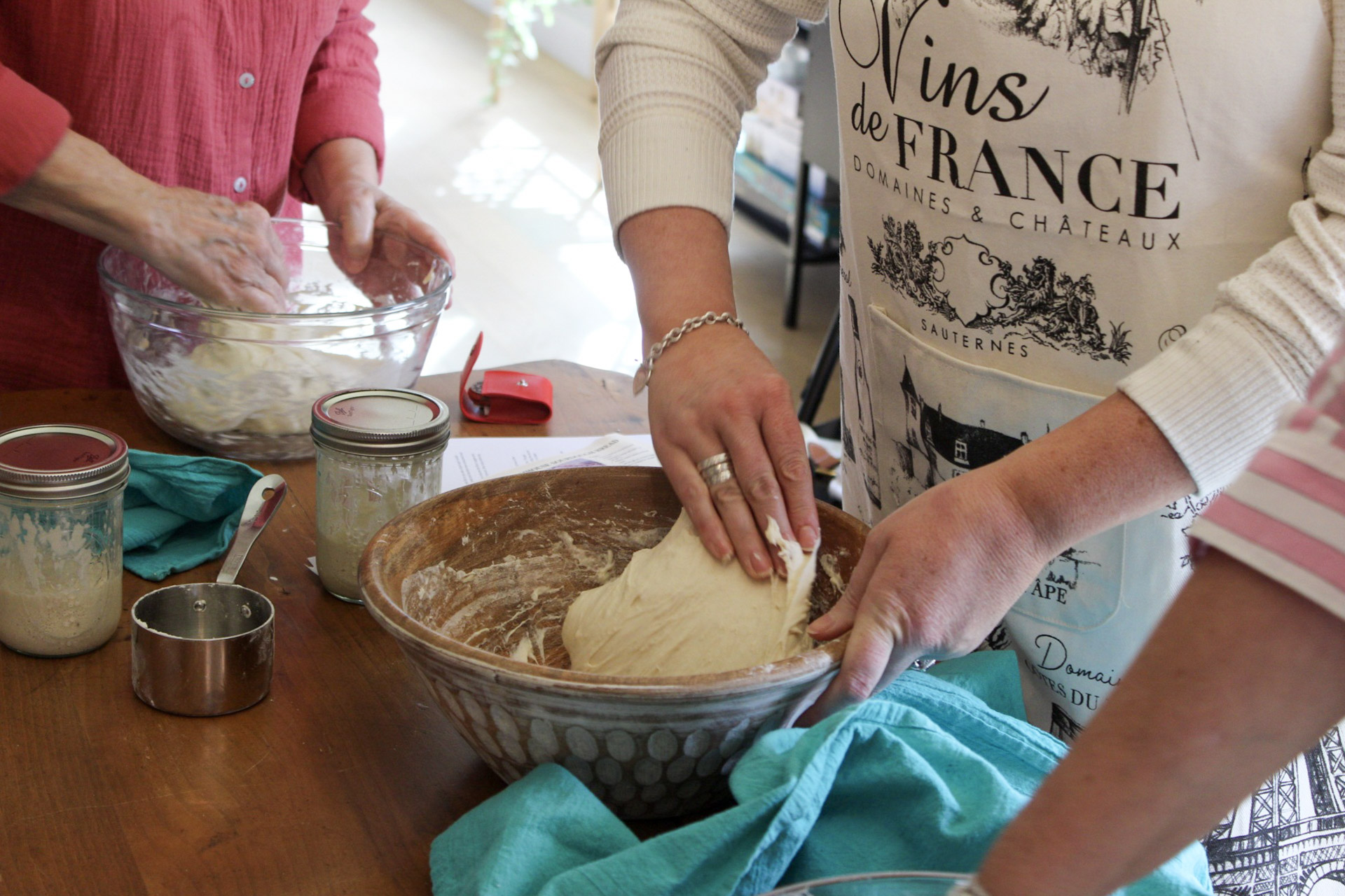 People kneading dough in a glass bowl on a wooden table, with baking ingredients and utensils nearby.