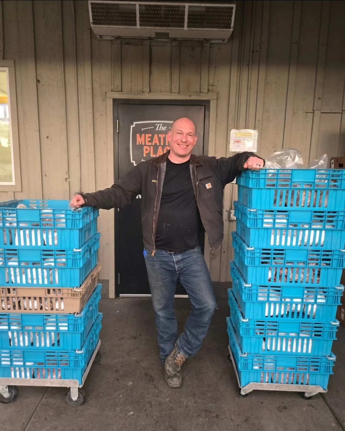 Man standing between two stacks of blue plastic crates, smiling, in front of a door with a sign, in an indoor setting.