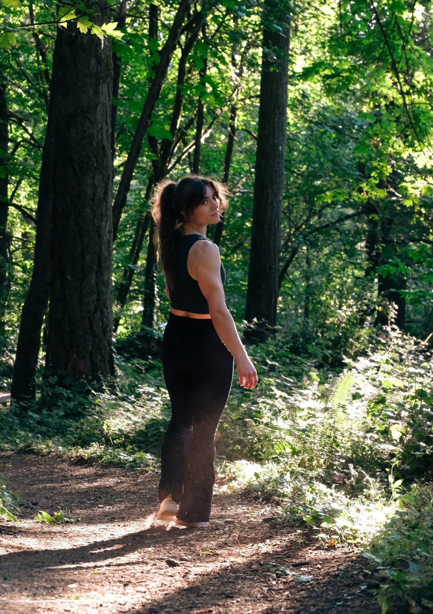 Woman standing on a forest trail surrounded by green trees and sunlight, facing sideways with her hair in a ponytail.