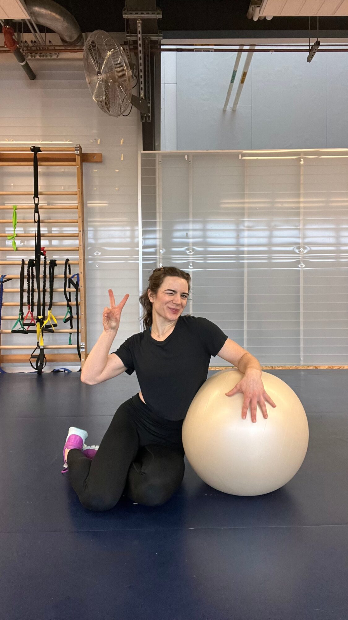 Person kneeling on floor with a large exercise ball, smiling and making a peace sign, in a gym setting.