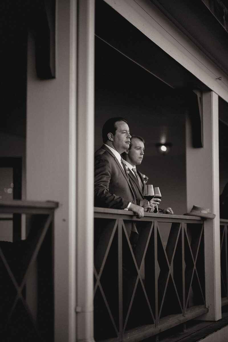 Two men in suits stand on a balcony, holding drinks, looking outward, with a dark background and a small light. Word count: 20