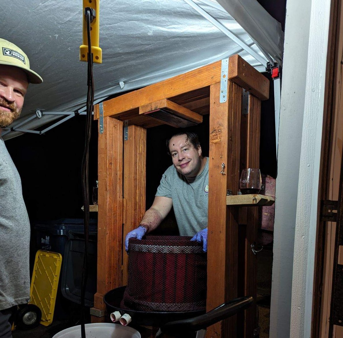 Two people working on a wooden structure inside a tent, one smiling and wearing gloves, the other partially visible.