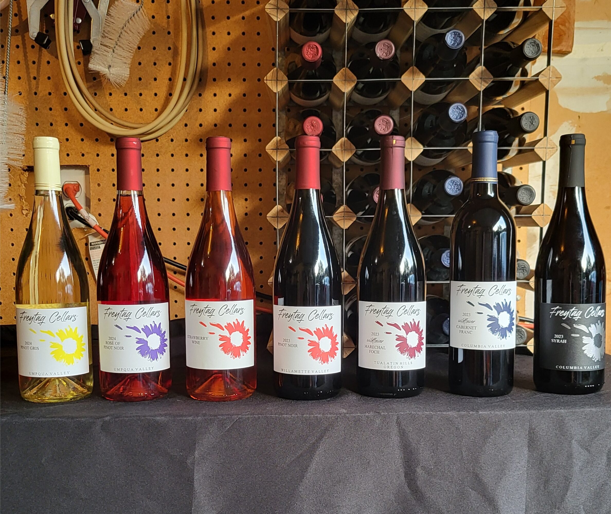 Six bottles of wine with colorful labels are displayed on a table, with a wine rack and tools in the background.