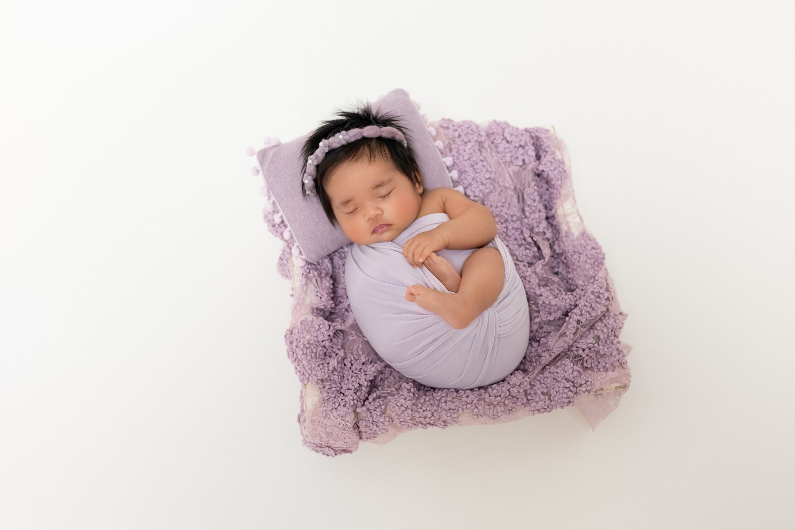 Baby sleeping on a purple blanket, wrapped in a light-colored cloth, on a white background.