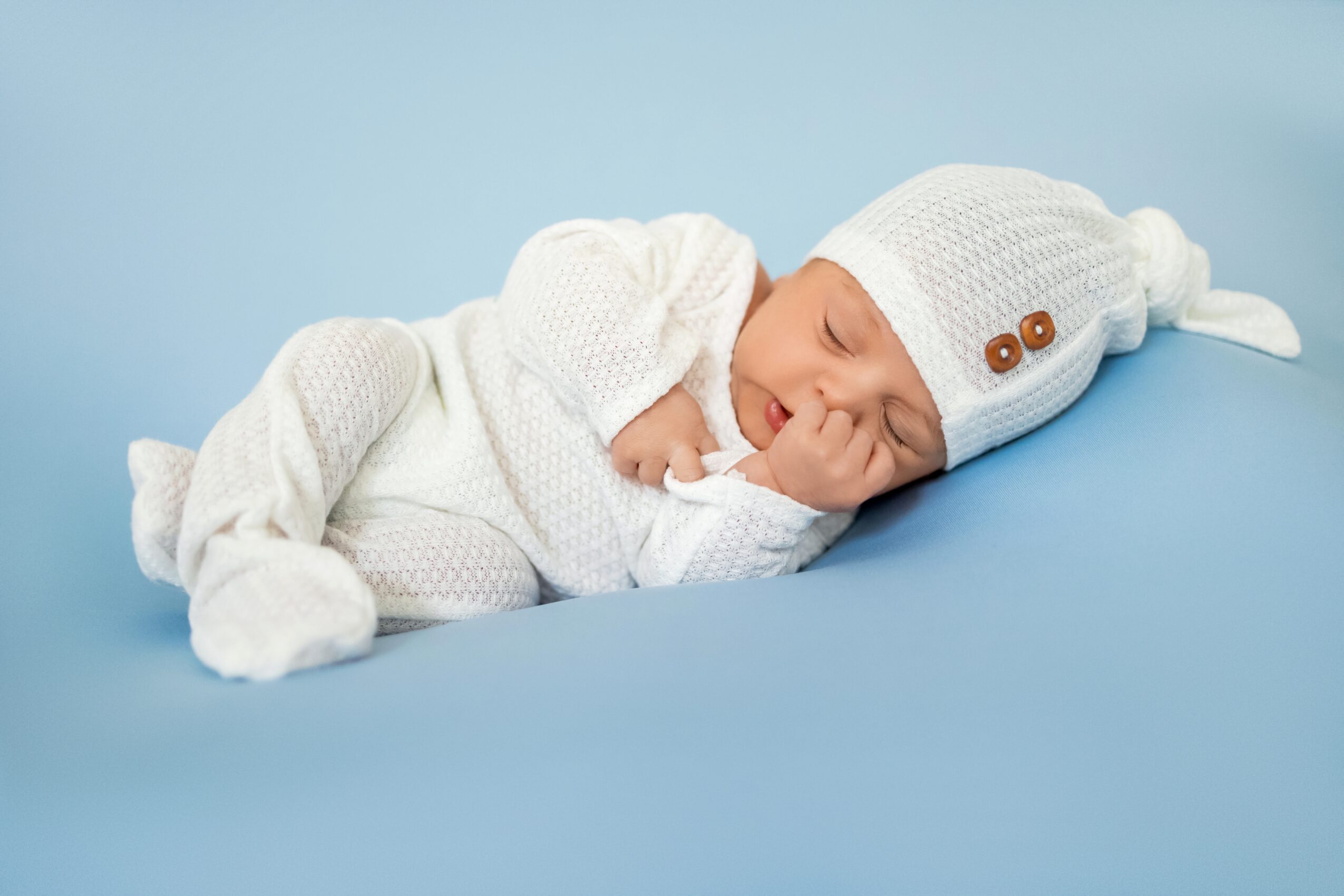Newborn baby lying on side, wearing a white knit hat and outfit, resting with hand near face, on light blue surface.