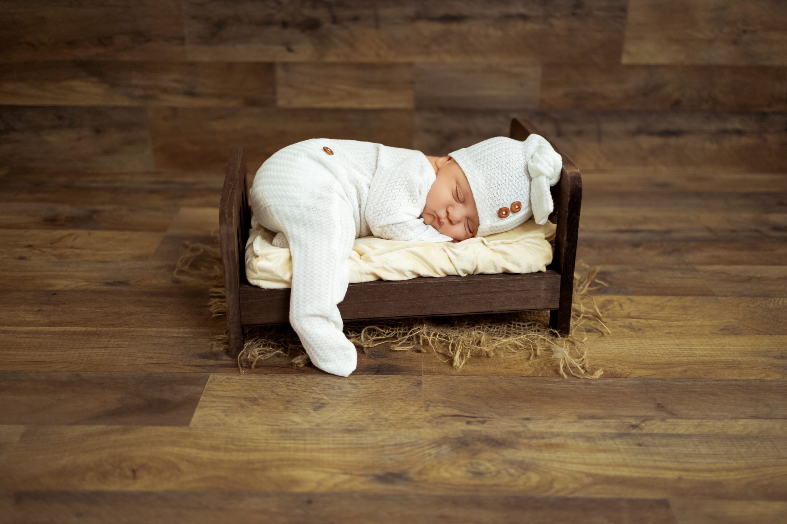 Baby dressed in white pajamas sleeping on a small wooden bed on a wooden floor.