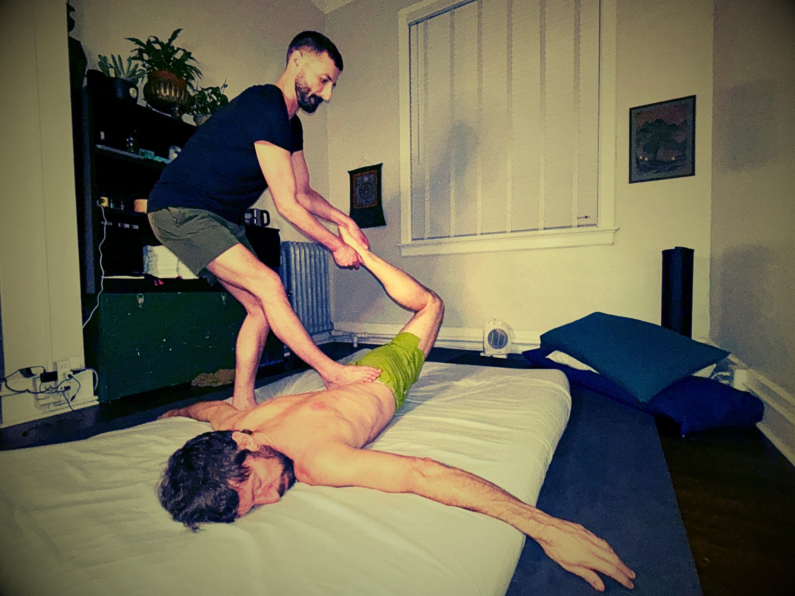Two men are on a bed, one lying down and the other standing, holding the other's arm. The room has a window, paintings, and furniture.