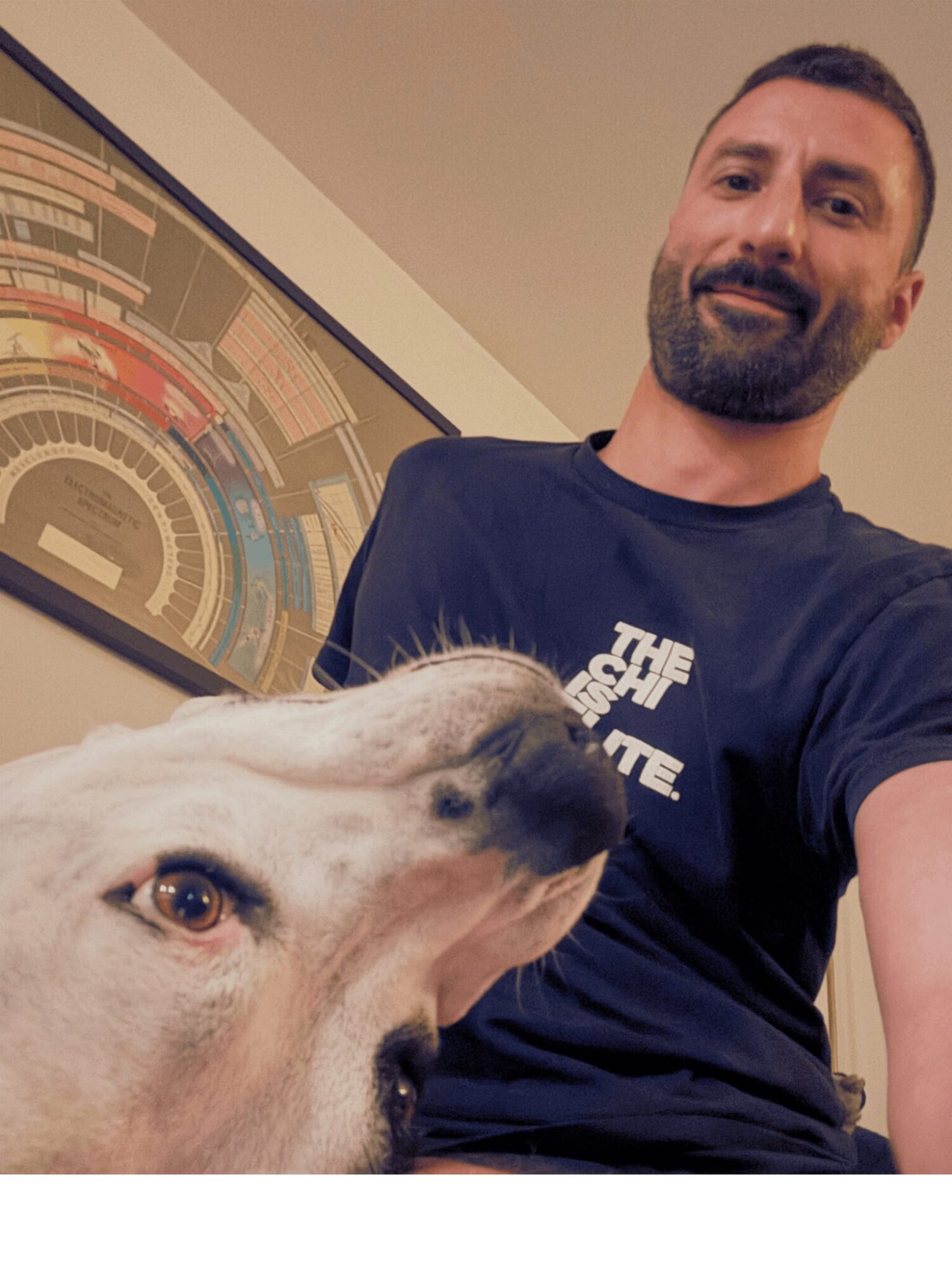 Man with beard smiling, taking a selfie with a large dog in front, colorful artwork on wall behind them.