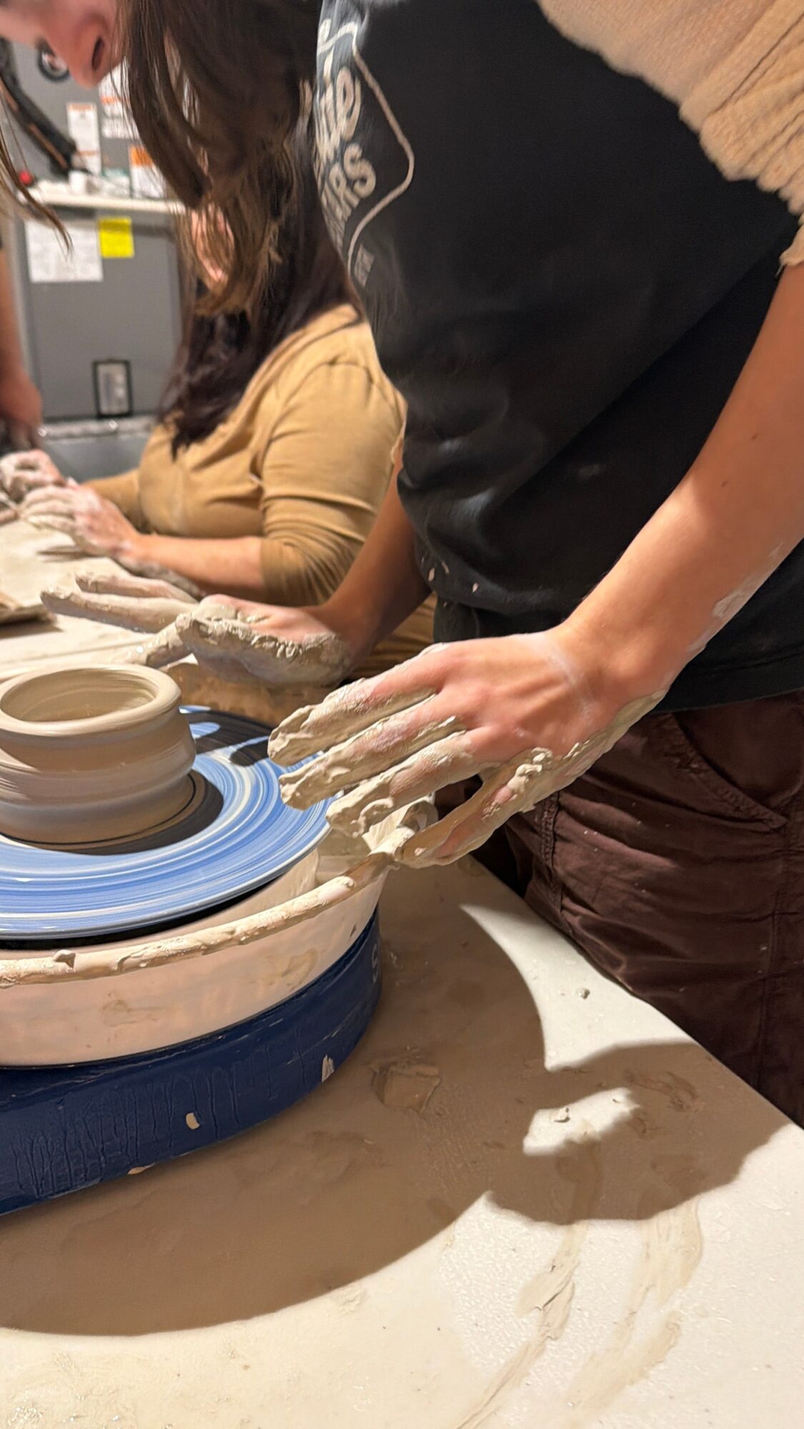 Person shaping clay on pottery wheel with hands covered in clay, others working nearby, workshop setting.