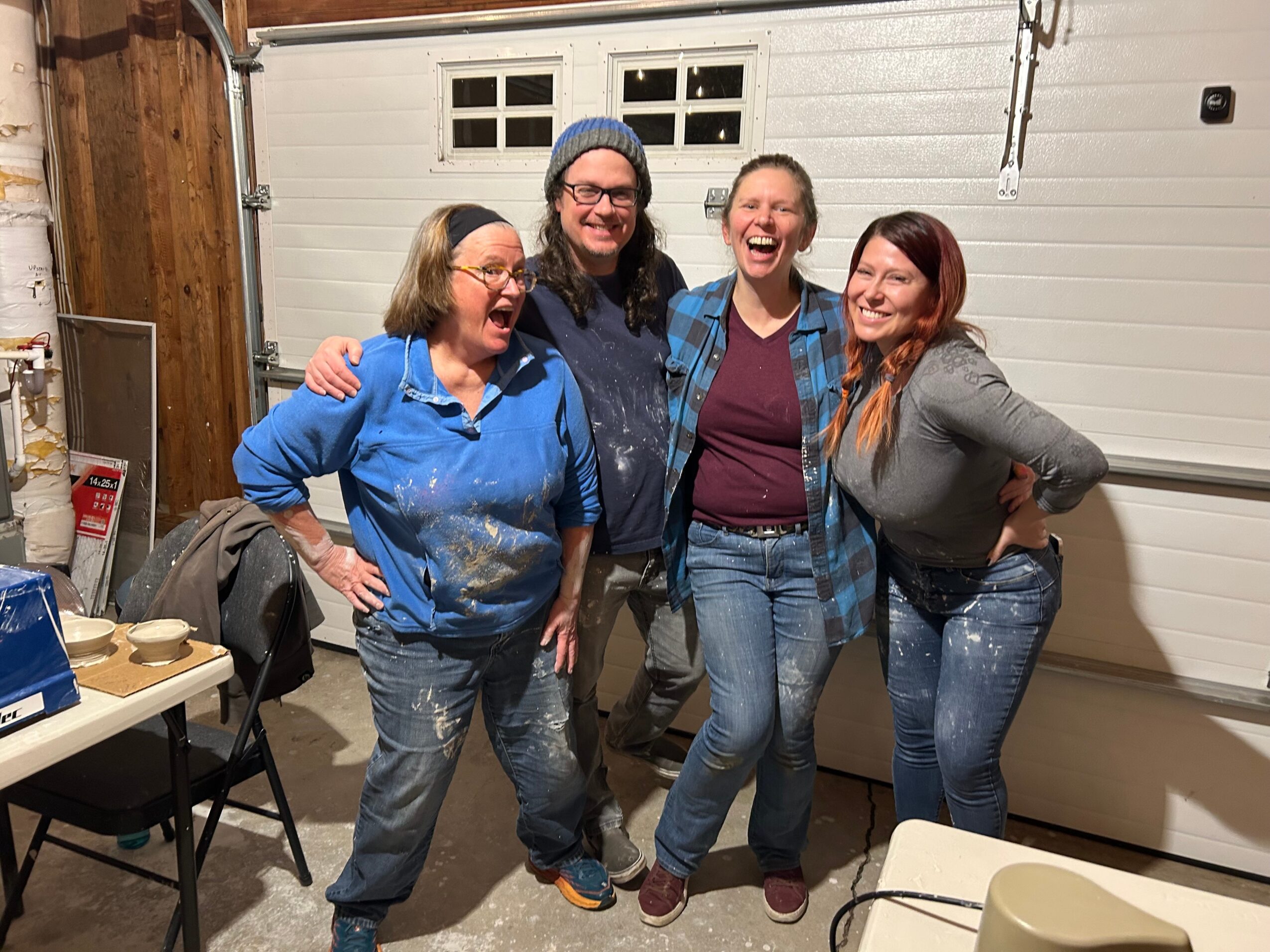 Four women smiling and posing together indoors, standing close with arms around each other, in front of a garage door.