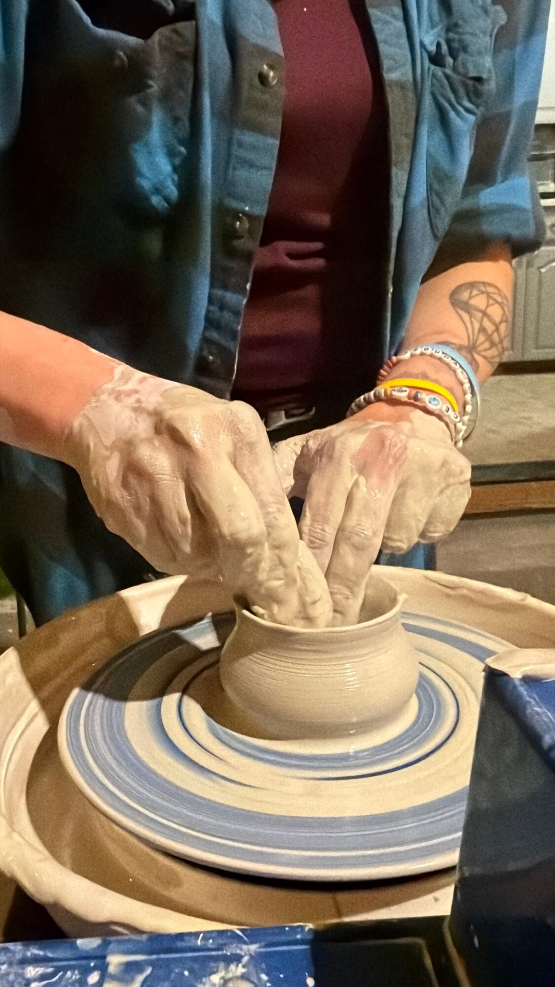 Person's hands shaping clay on a pottery wheel, wearing bracelets and a tattooed arm, with a focused posture.