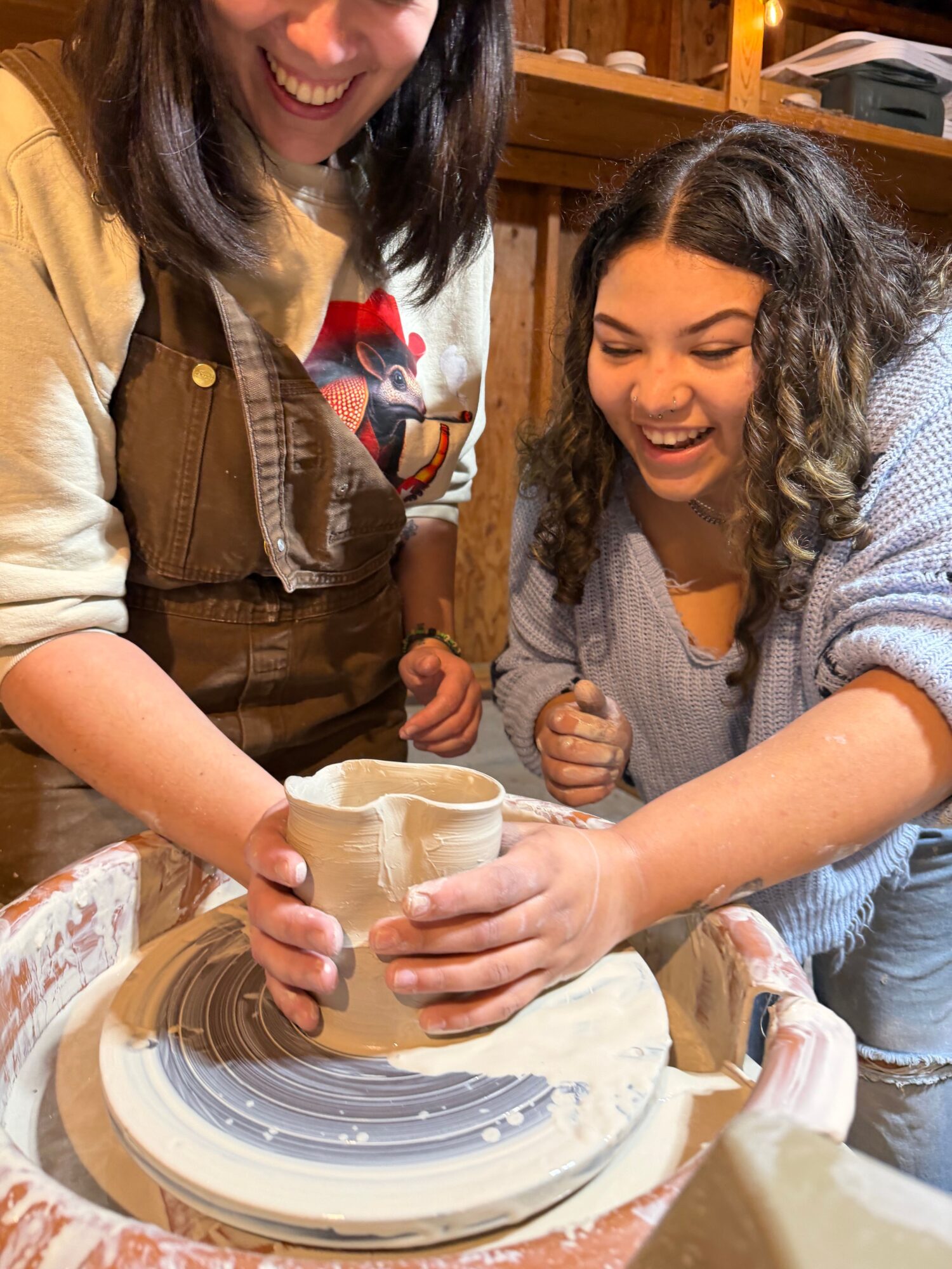 Two women are working together on a pottery wheel, shaping a clay object. They are smiling and focused on their task.