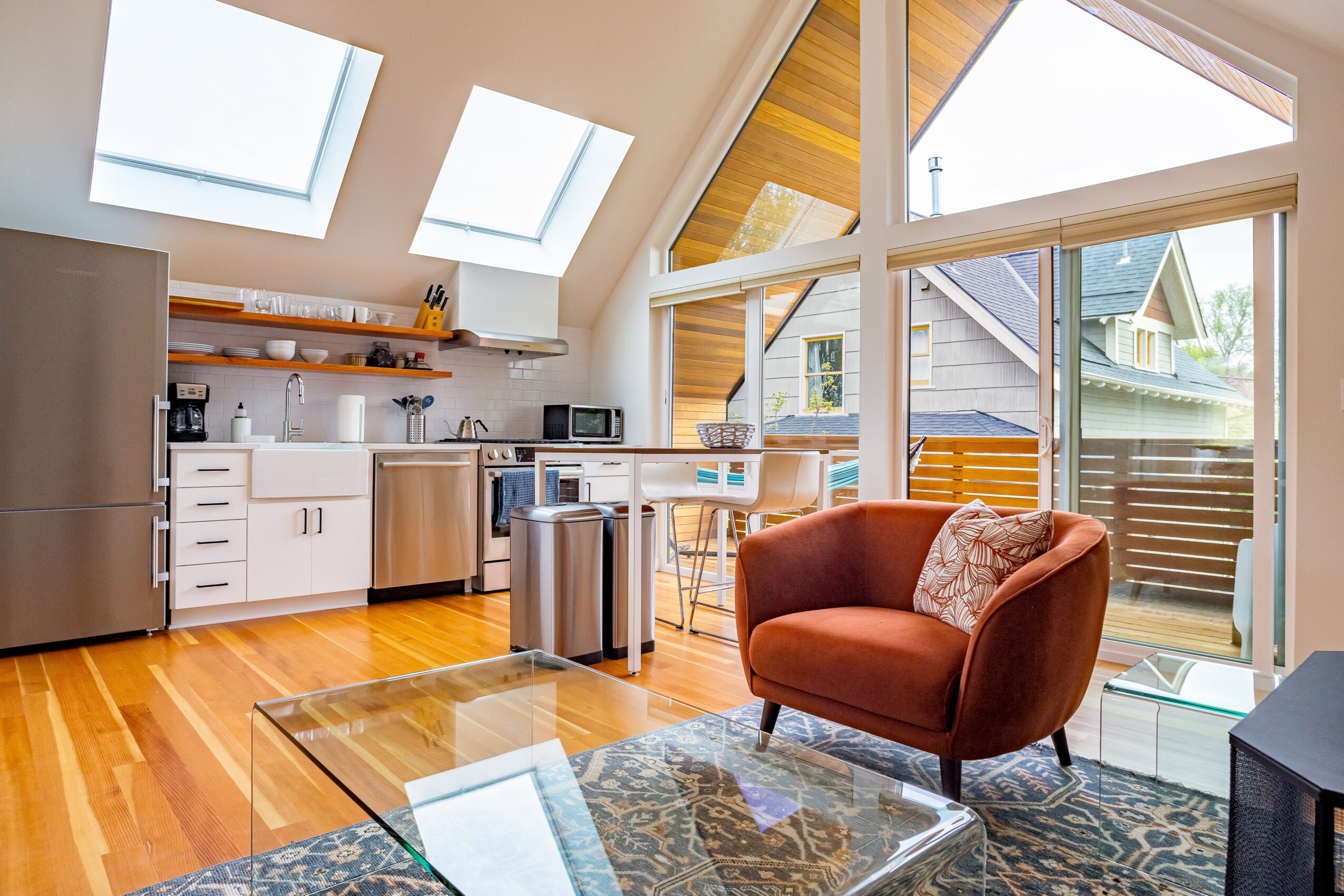 Living area with a red armchair, glass coffee table, and large windows with a view of neighboring houses.