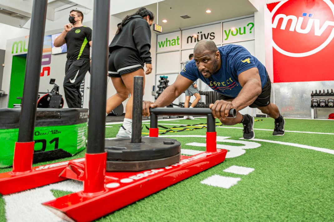 Man doing push-up with sled on artificial turf, two women in background, gym setting with signs and equipment.