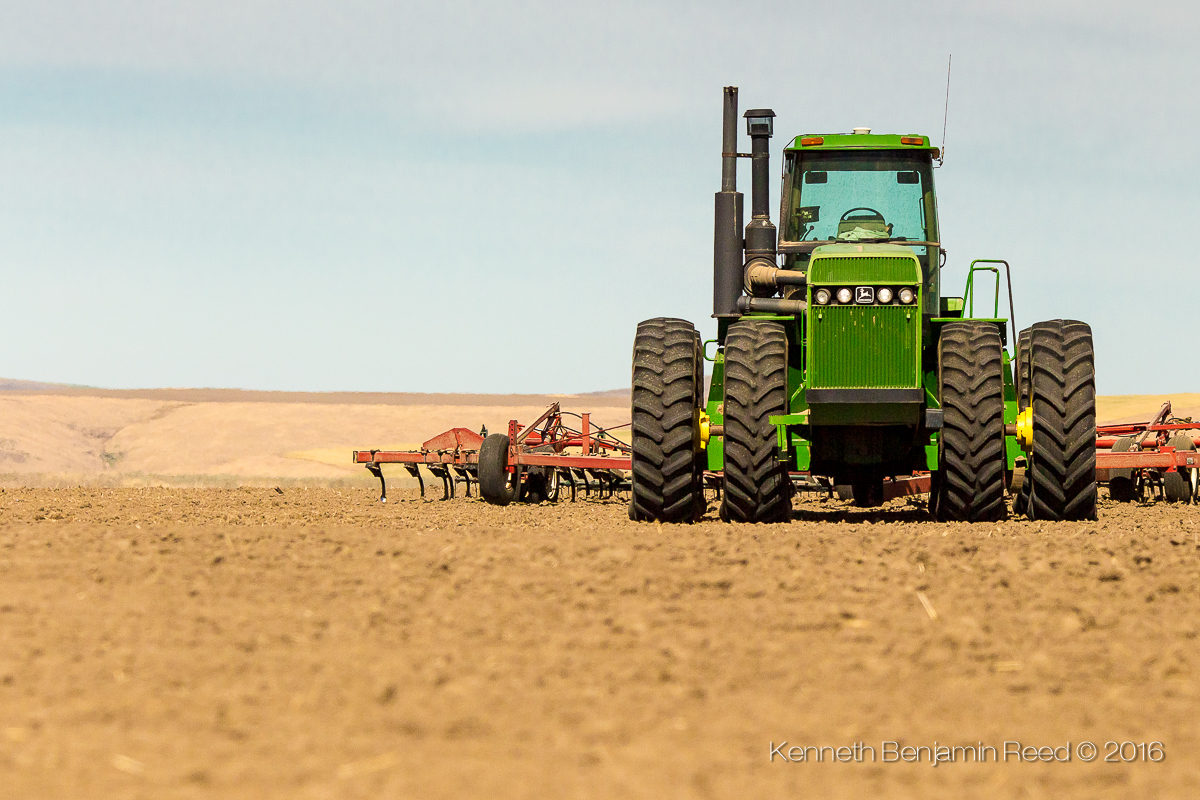 Green tractor on a farm field with farming equipment in the background, under a clear sky.
