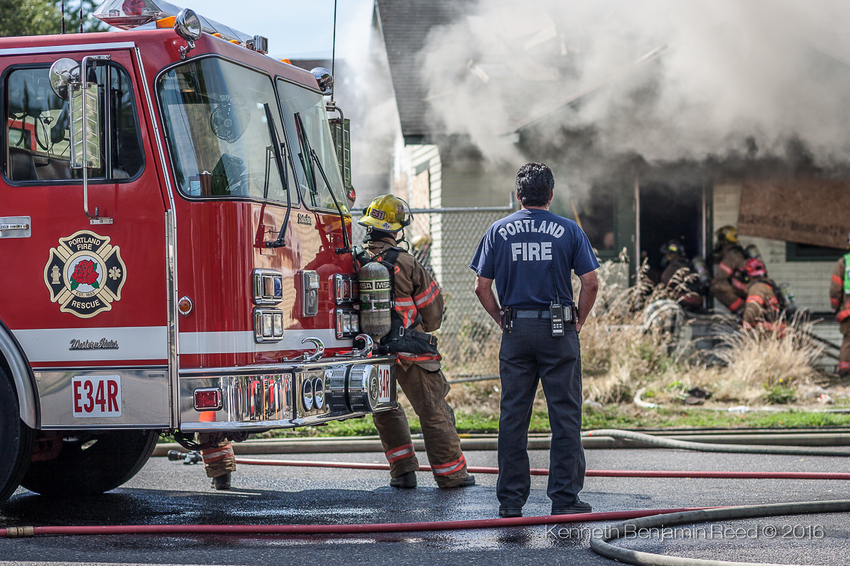 Firefighters and a person in a Portland Fire Department uniform respond to a fire with smoke billowing from a building, fire truck nearby.