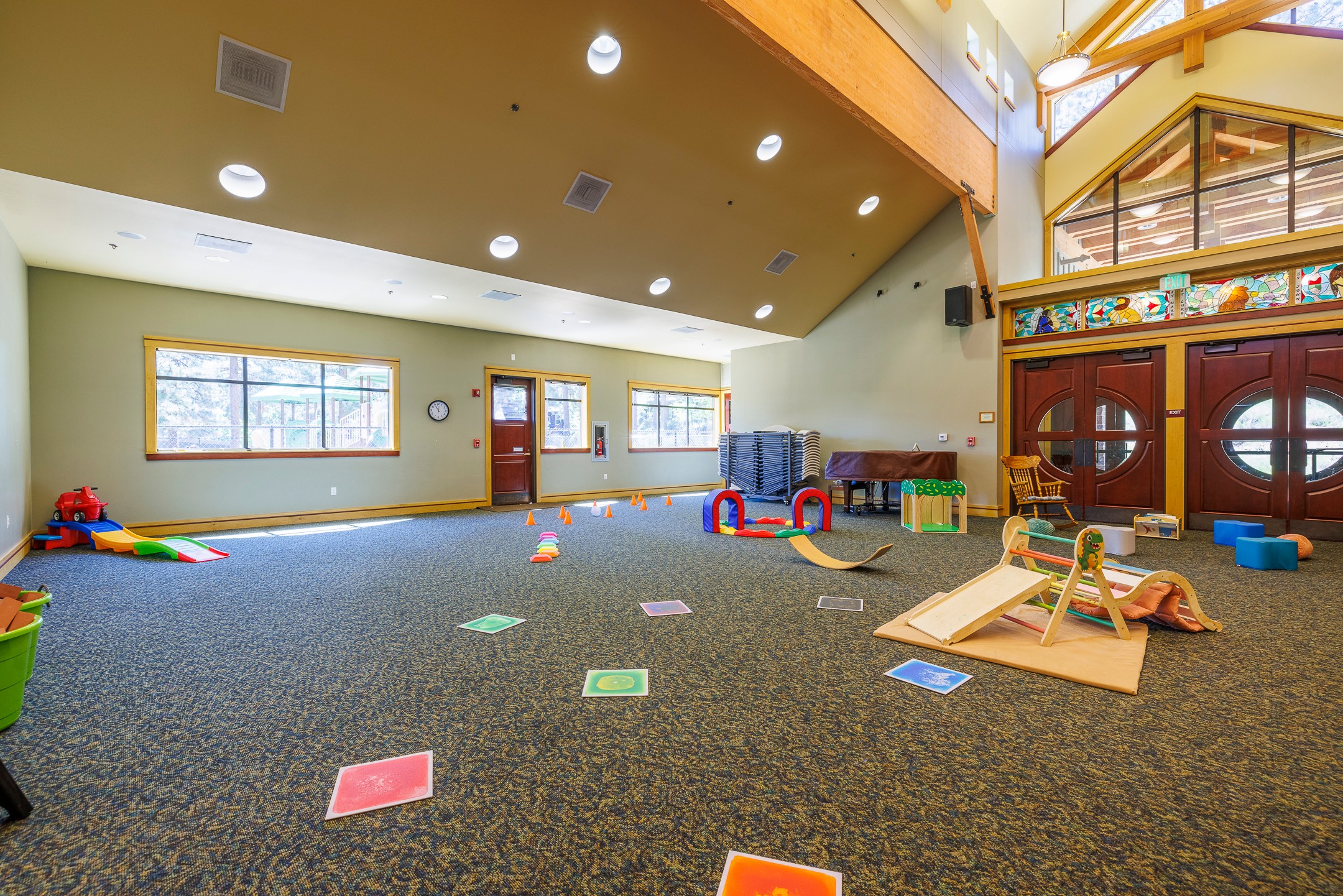 Indoor playroom with colorful toys, mats, and a wooden structure, bright windows, and a high ceiling with lights.