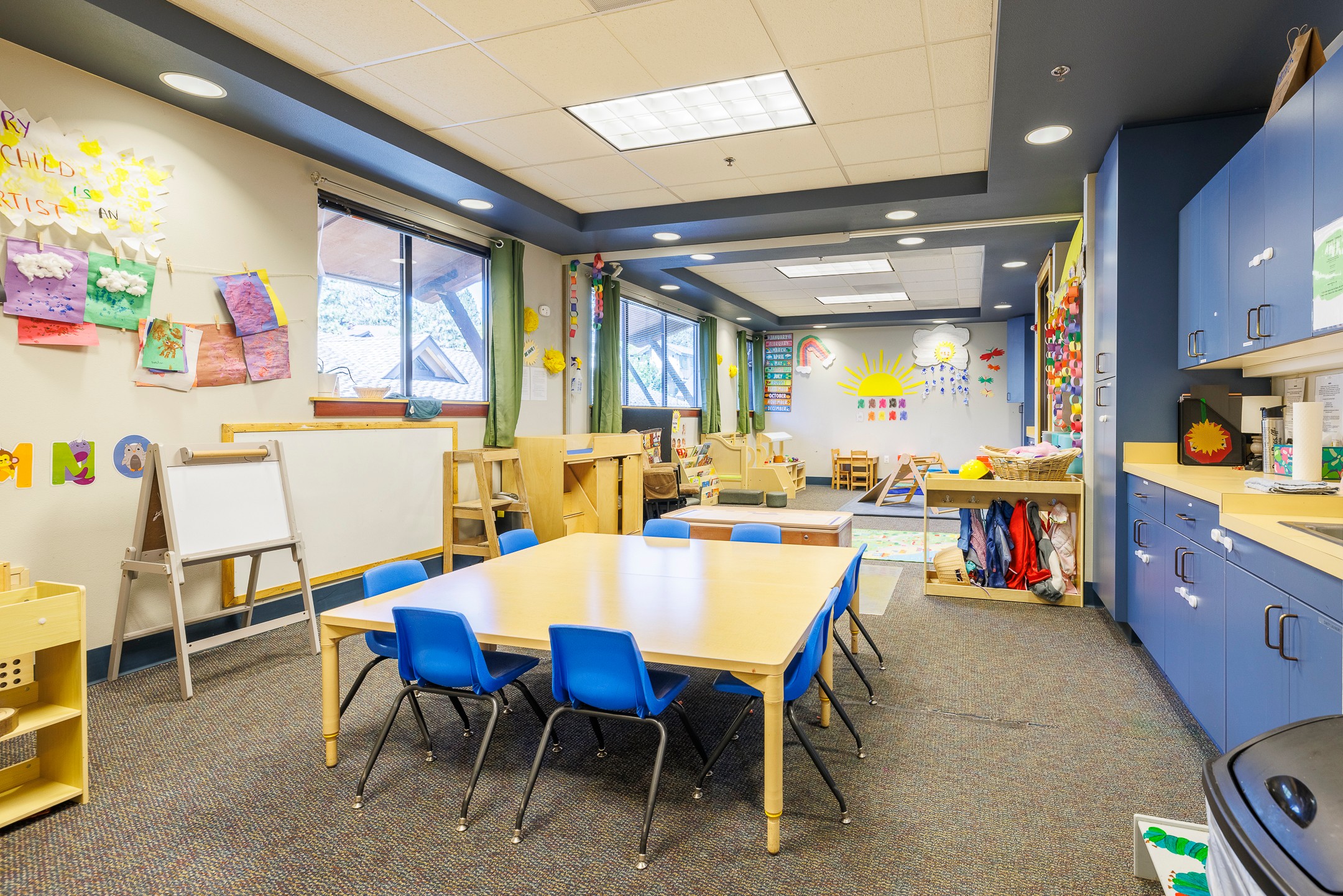 Classroom with tables, chairs, and colorful decorations, bright windows, and storage cabinets, designed for young children.