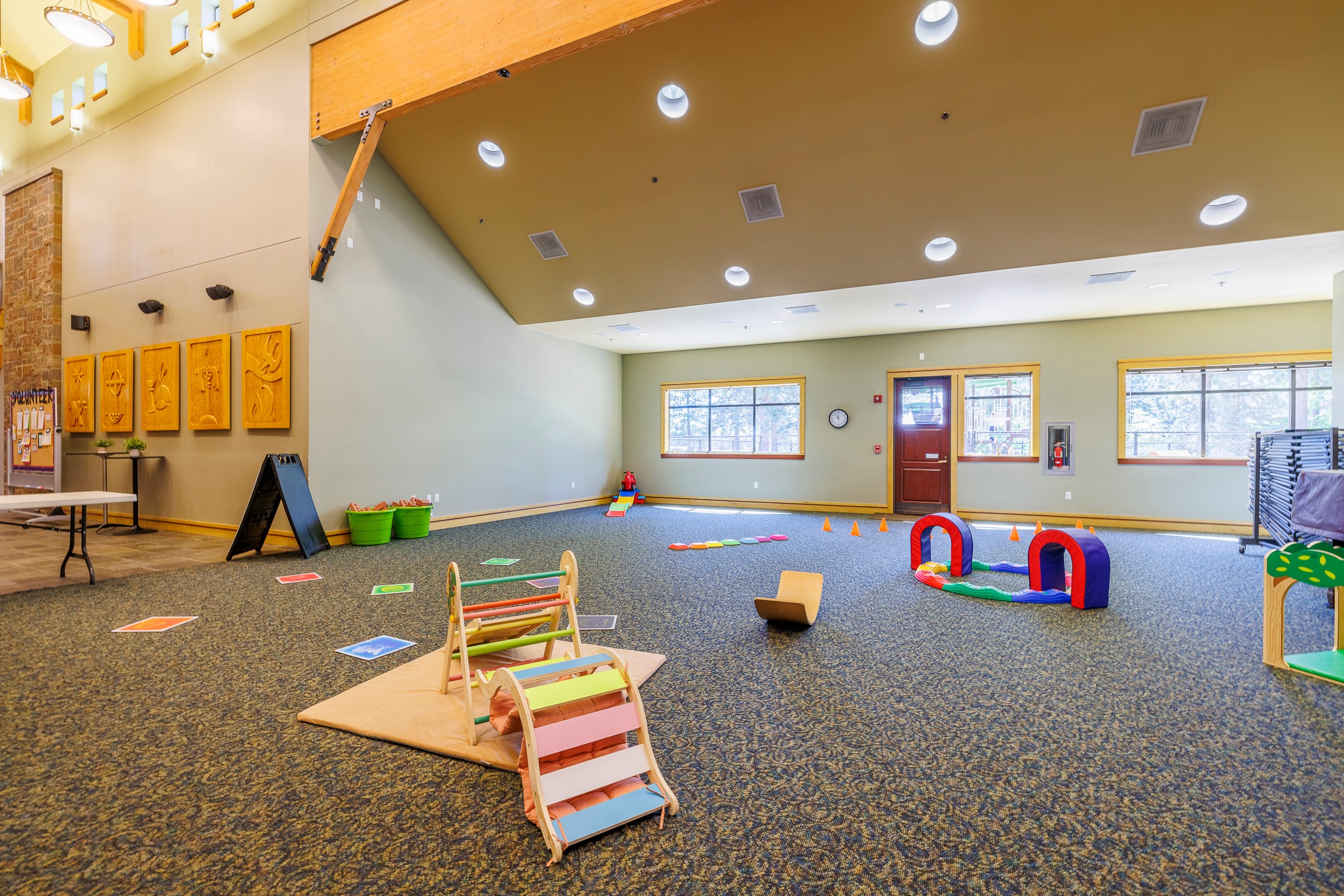 Indoor playroom with colorful equipment, windows, and a carpeted floor, viewed from the side with a sloped ceiling.