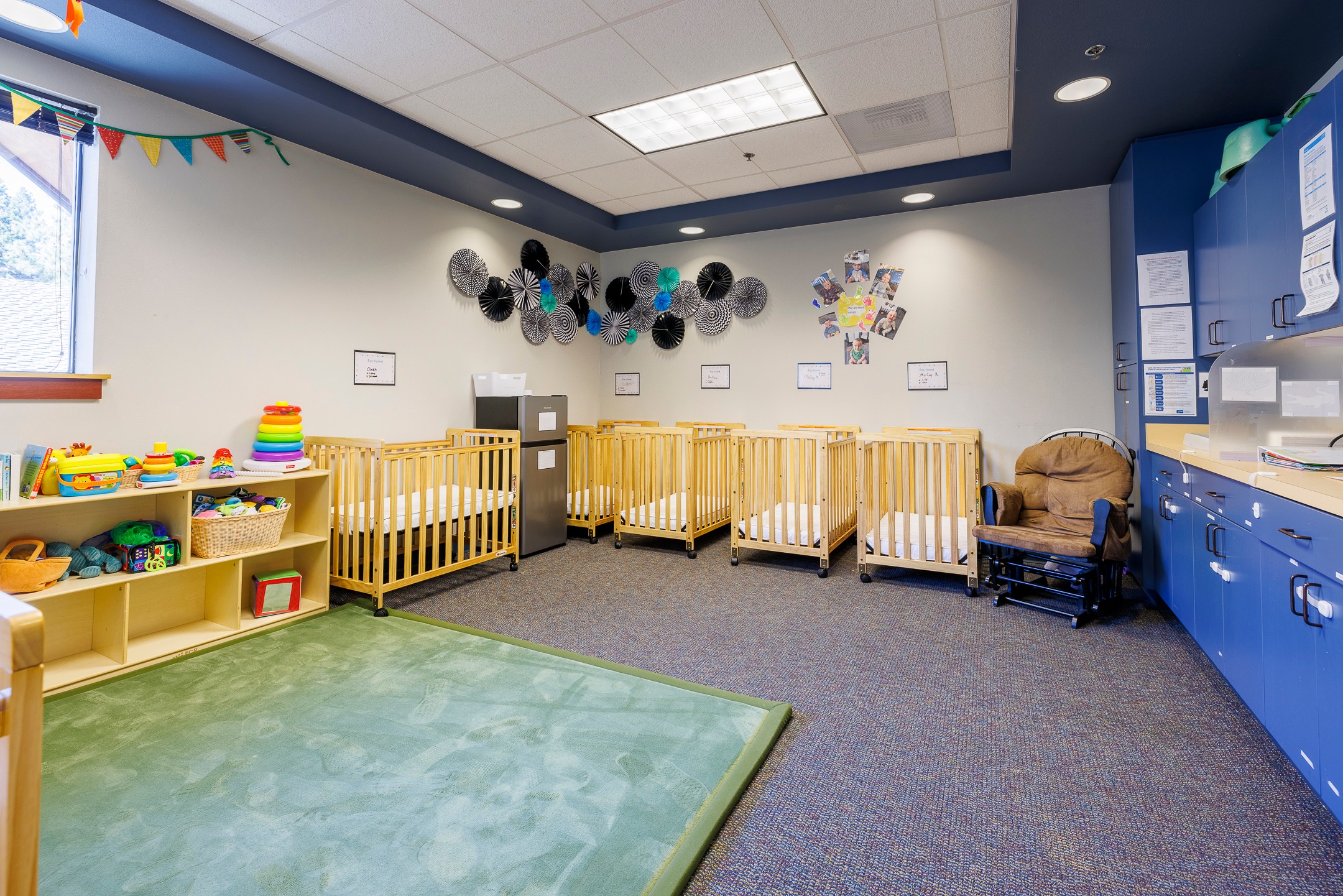 Nursery room with cribs, toys, and colorful decorations, including a green play mat and blue storage cabinets.