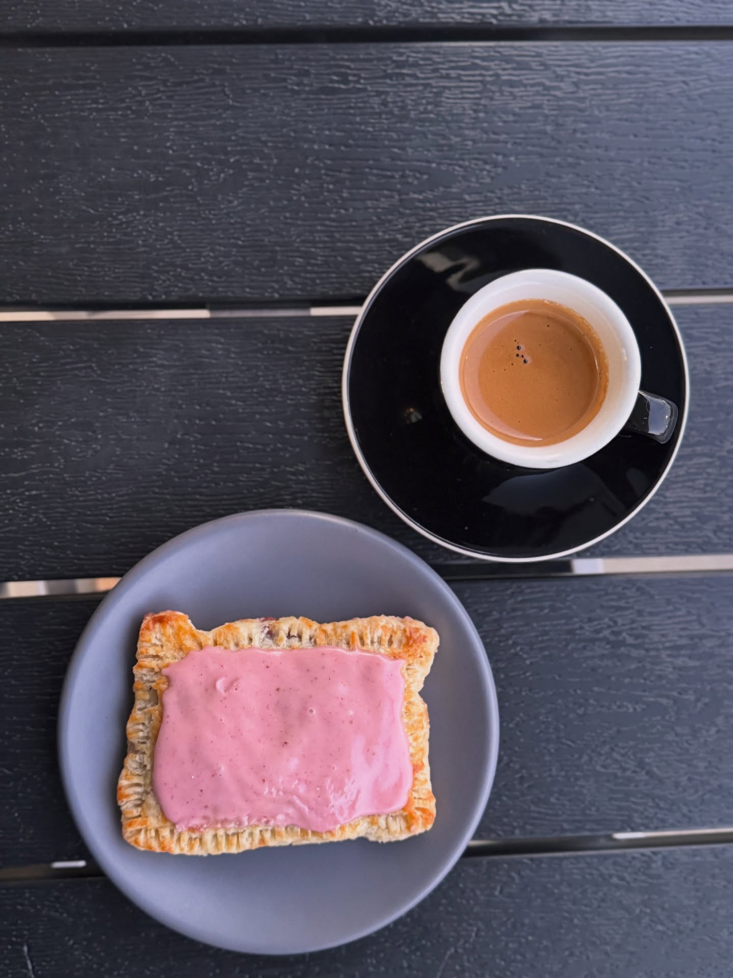 A cup of coffee on a black saucer and a slice of bread with pink spread on a gray plate, on a dark wooden surface.
