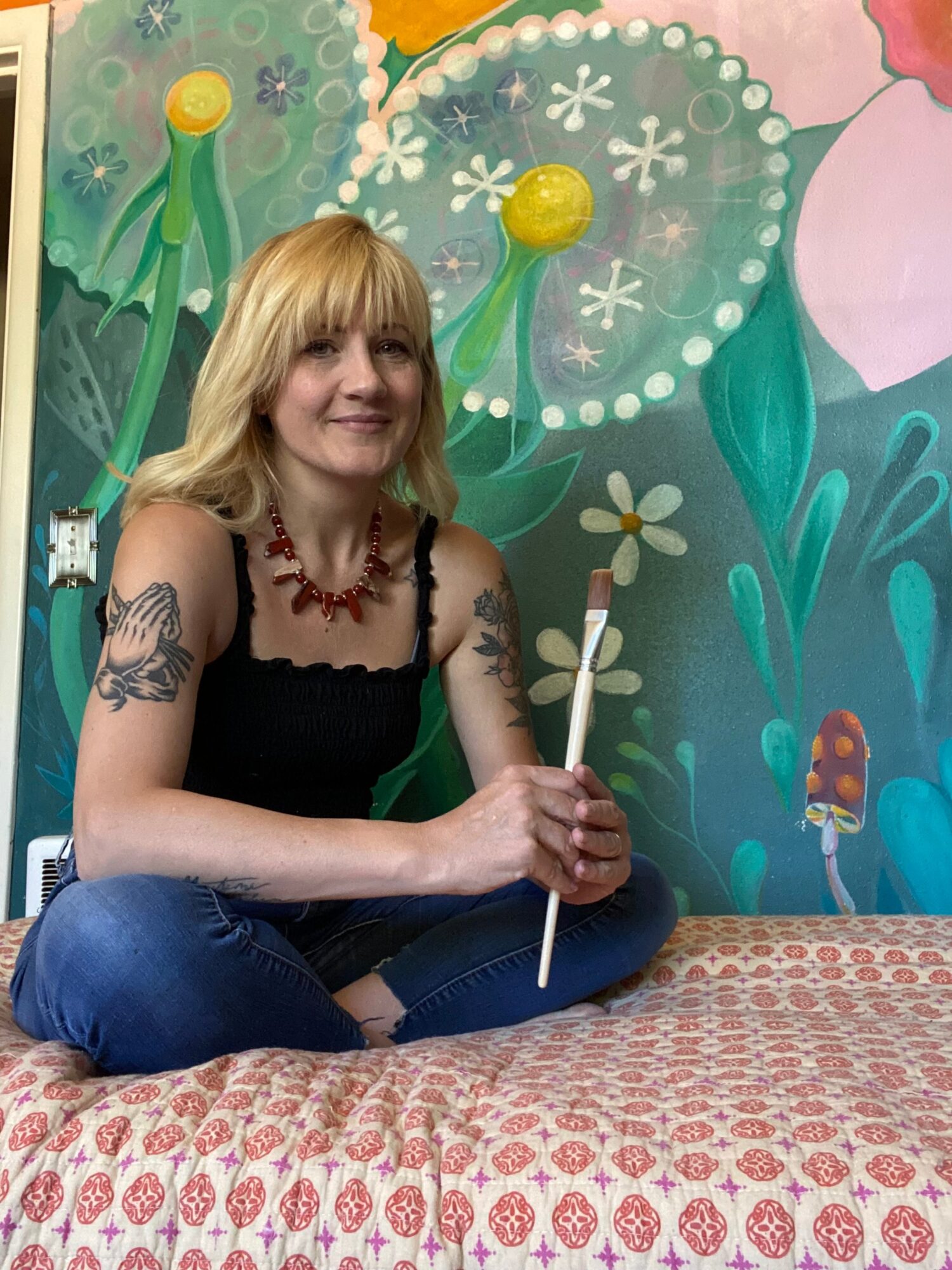 Woman with blonde hair, tattoos, and red necklace sitting cross-legged on a bed, holding a white stick, in front of a colorful floral mural.