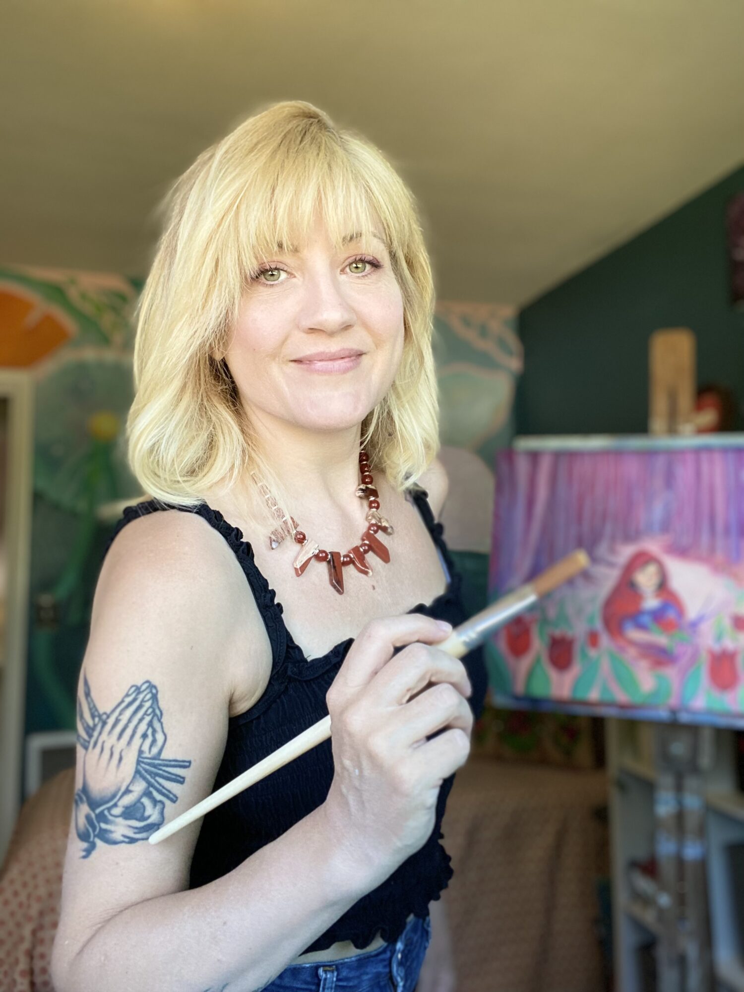 Woman with blonde hair holding a paintbrush, standing in front of a colorful painting, smiling.